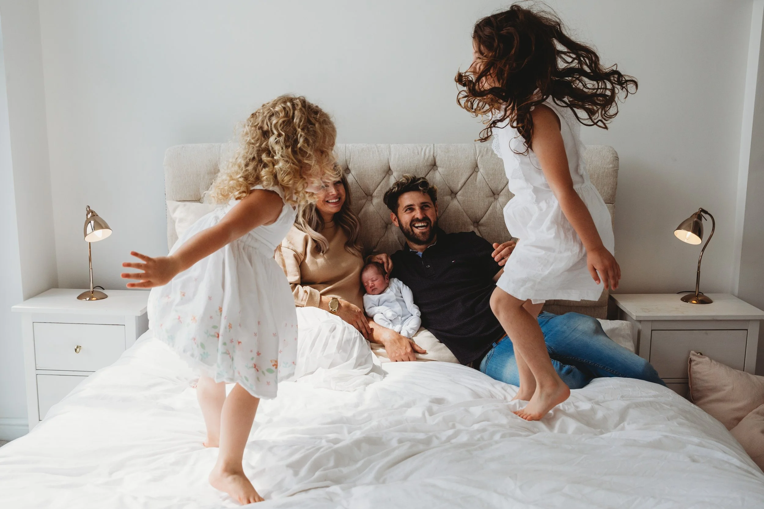Family enjoying a playful moment on a bed in a bedroom with white walls and two bedside lamps.