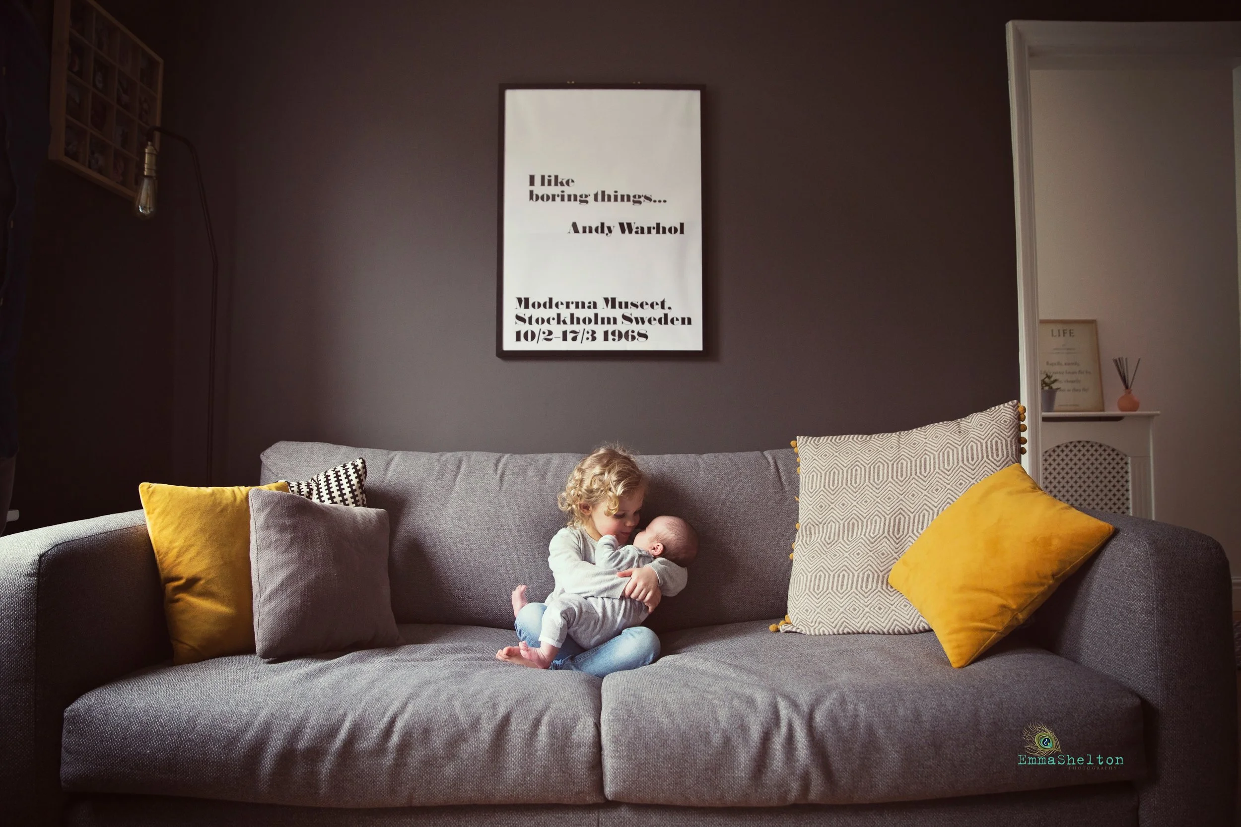 A living room with a large gray sofa decorated with yellow and patterned pillows. Two young children are sitting on the sofa, one holding the other. The background features a dark wall with a white framed poster about an art exhibition, and a partial