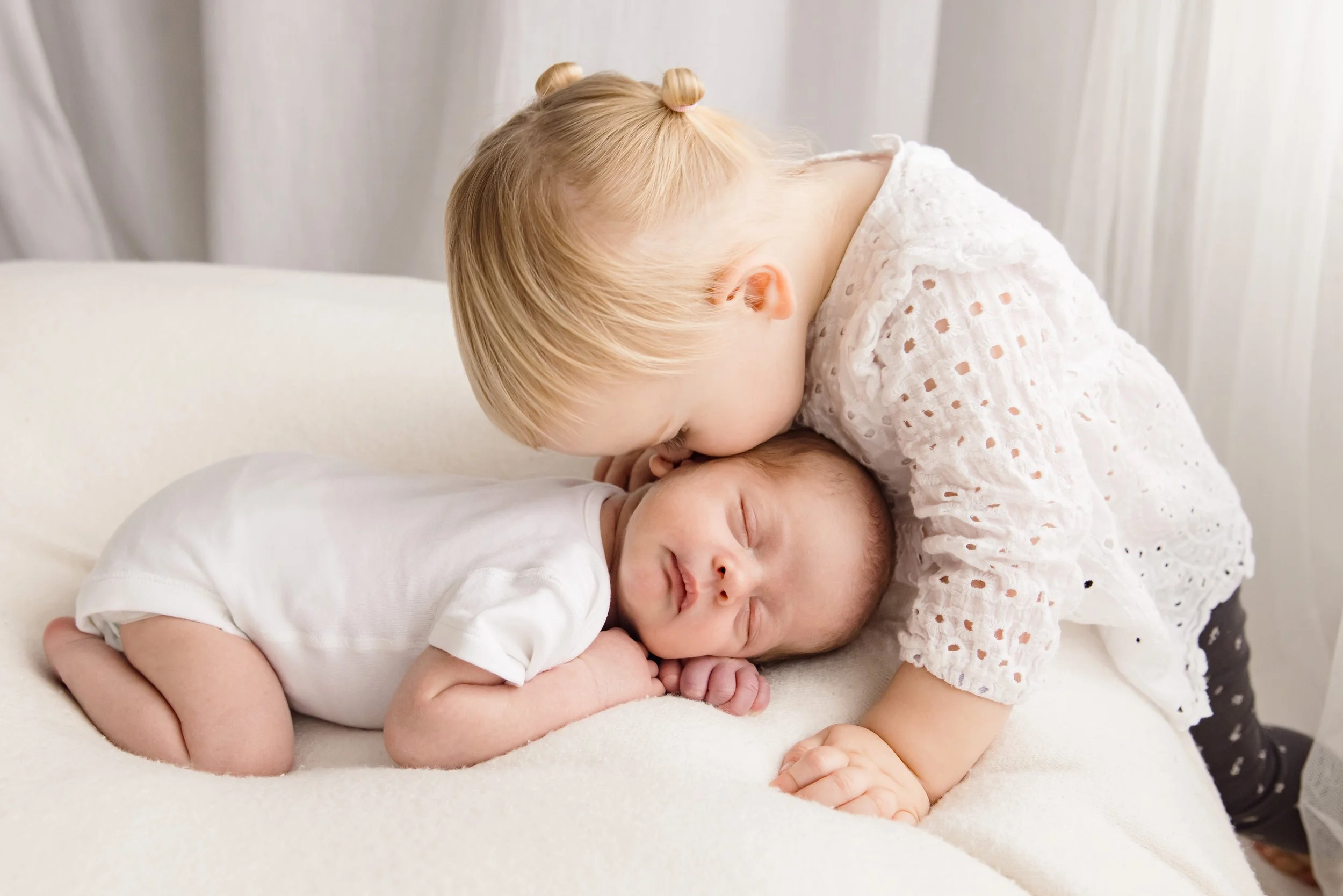 A young girl with blonde hair styled in two buns kissing a sleeping baby on the forehead. The baby is lying on a bed with cream-colored sheets, wearing a white onesie. The girl is wearing a white eyelet dress, and the background features light-colore