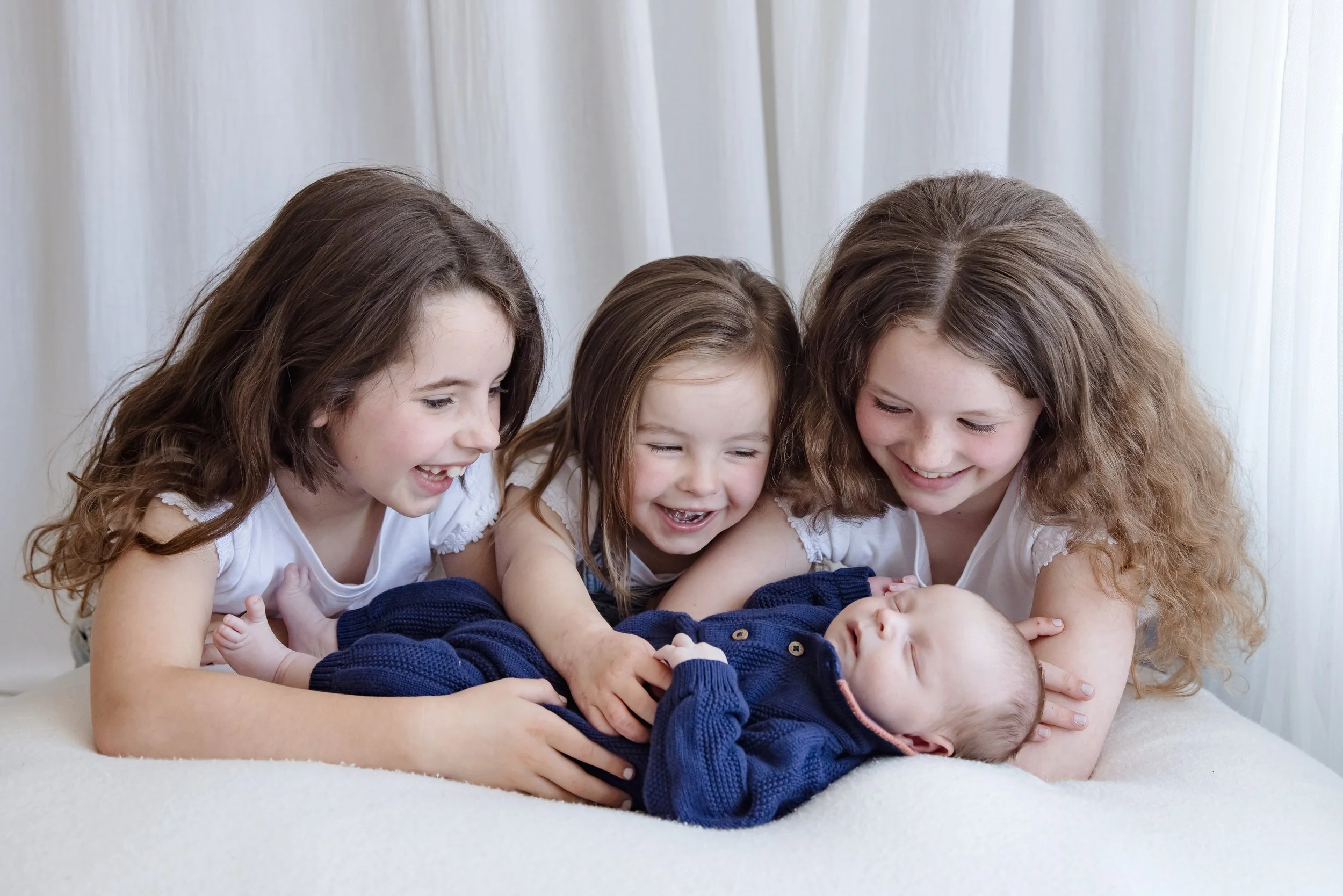Four young girls with long hair are smiling and playing with a baby lying on a blanket, all indoors in front of white curtains.