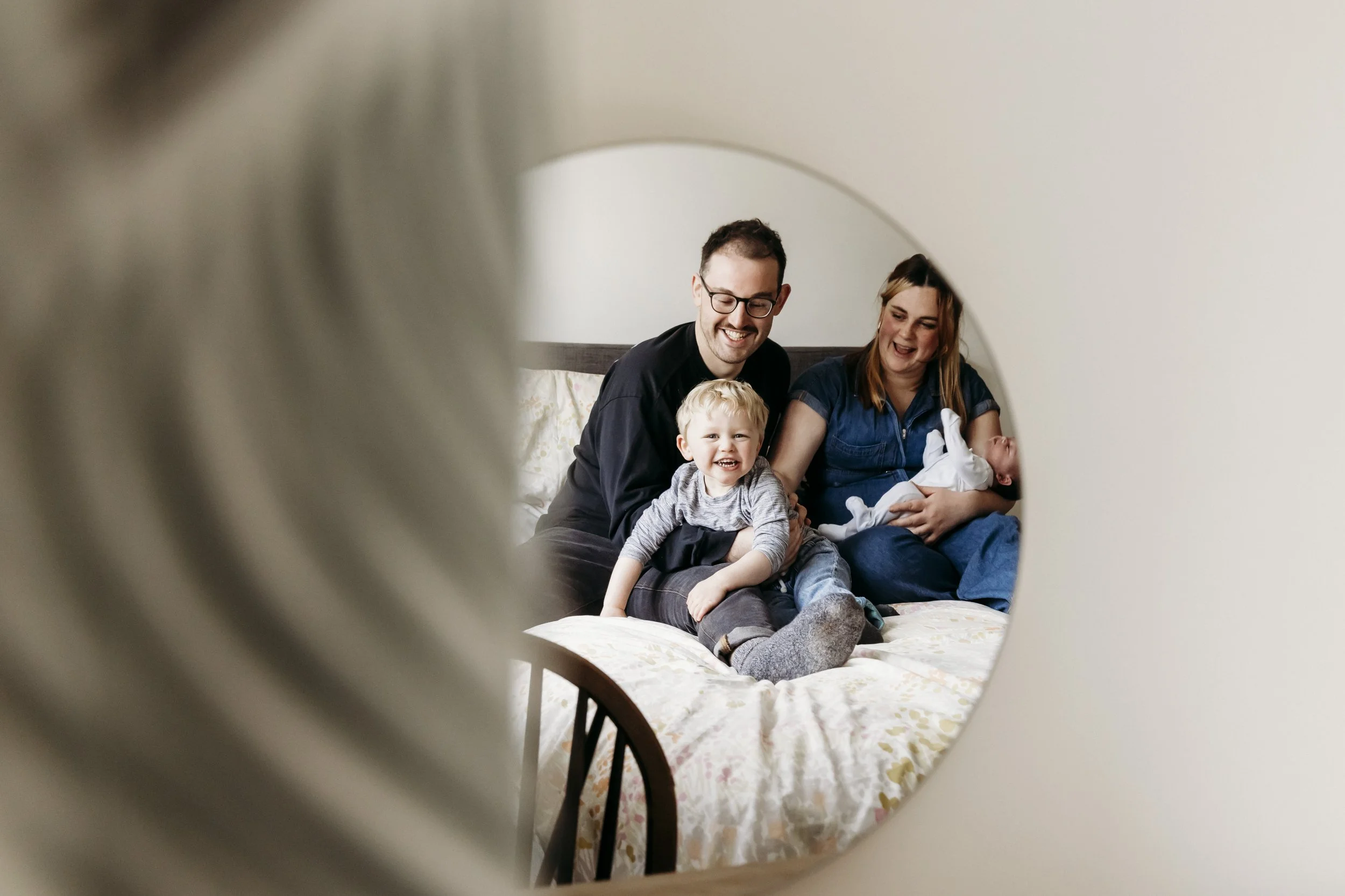 Family sitting on bed, smiling and laughing, as viewed through a round mirror.