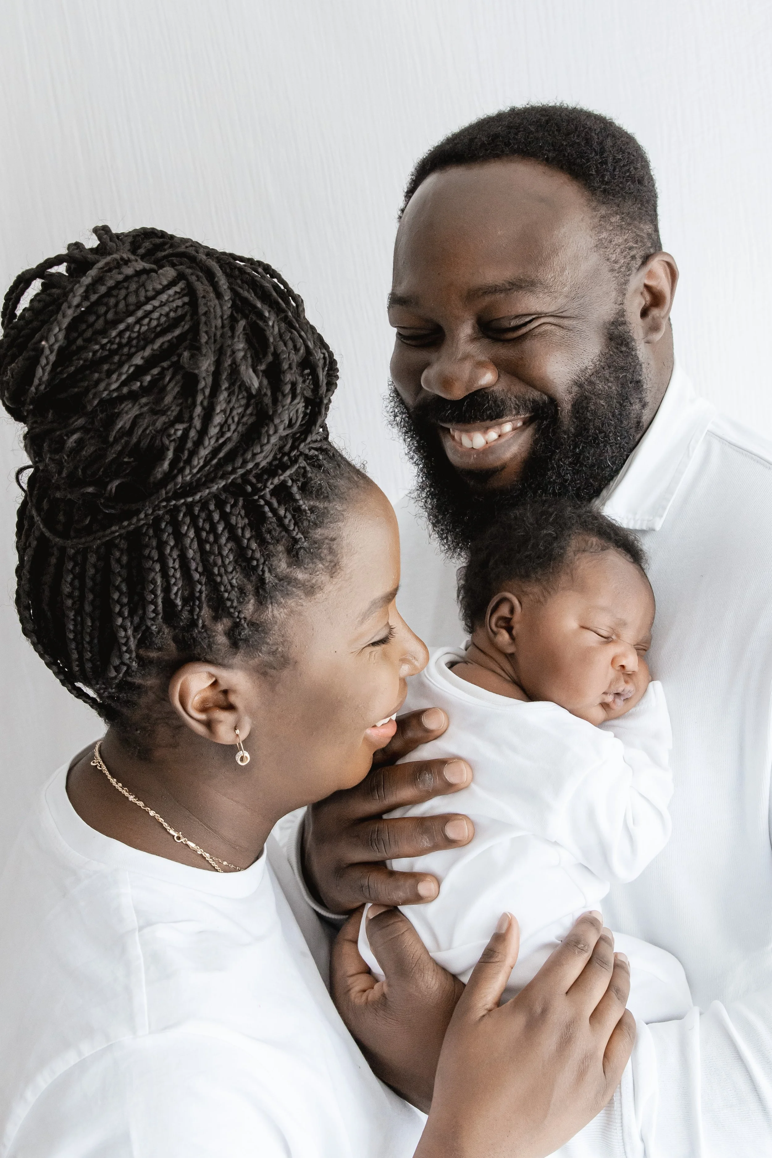 A smiling man holding a sleeping baby, a woman looking at the baby, all dressed in white against a plain white background.
