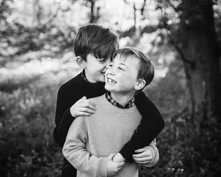 Two young boys sharing a happy moment outdoors, with one boy hugging the other and both smiling.
