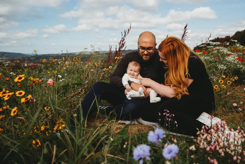 A family with a baby sitting on grass in a field of colorful flowers under a partly cloudy sky.
