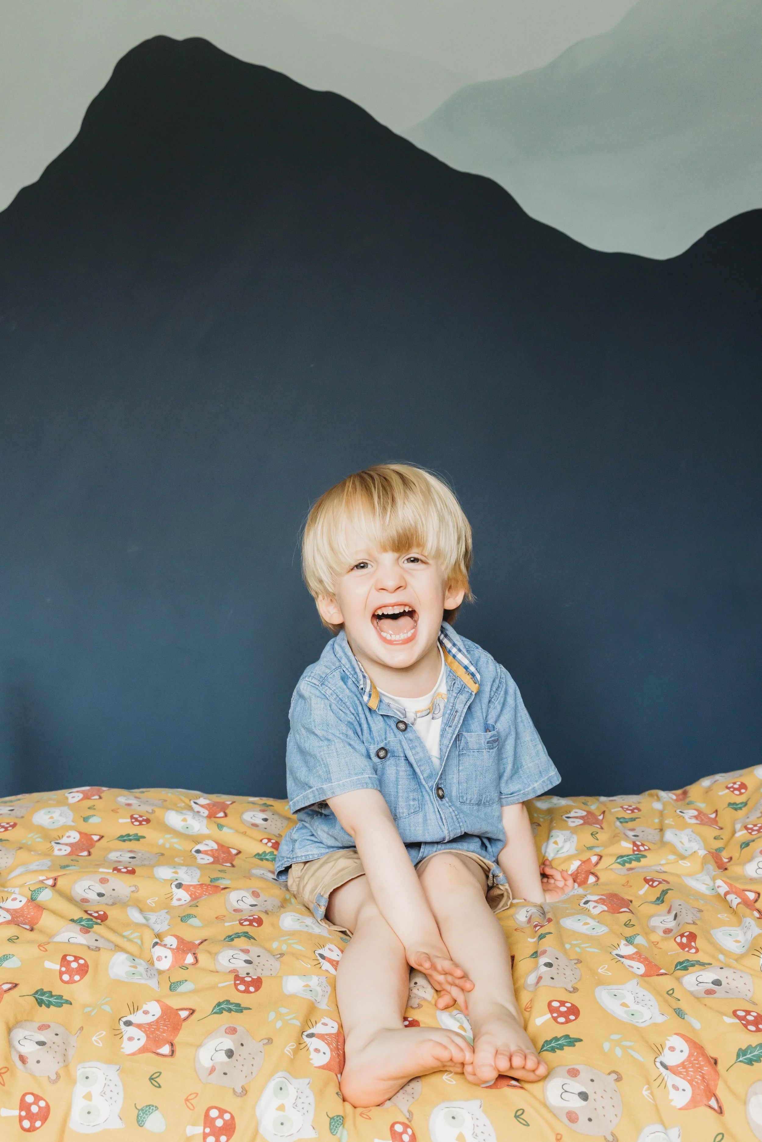 A young boy with blonde hair sitting on a brightly colored blanket with woodland creatures, in front of a painted mountain landscape wall. The boy is smiling and has his mouth open, wearing a denim shirt and beige shorts.