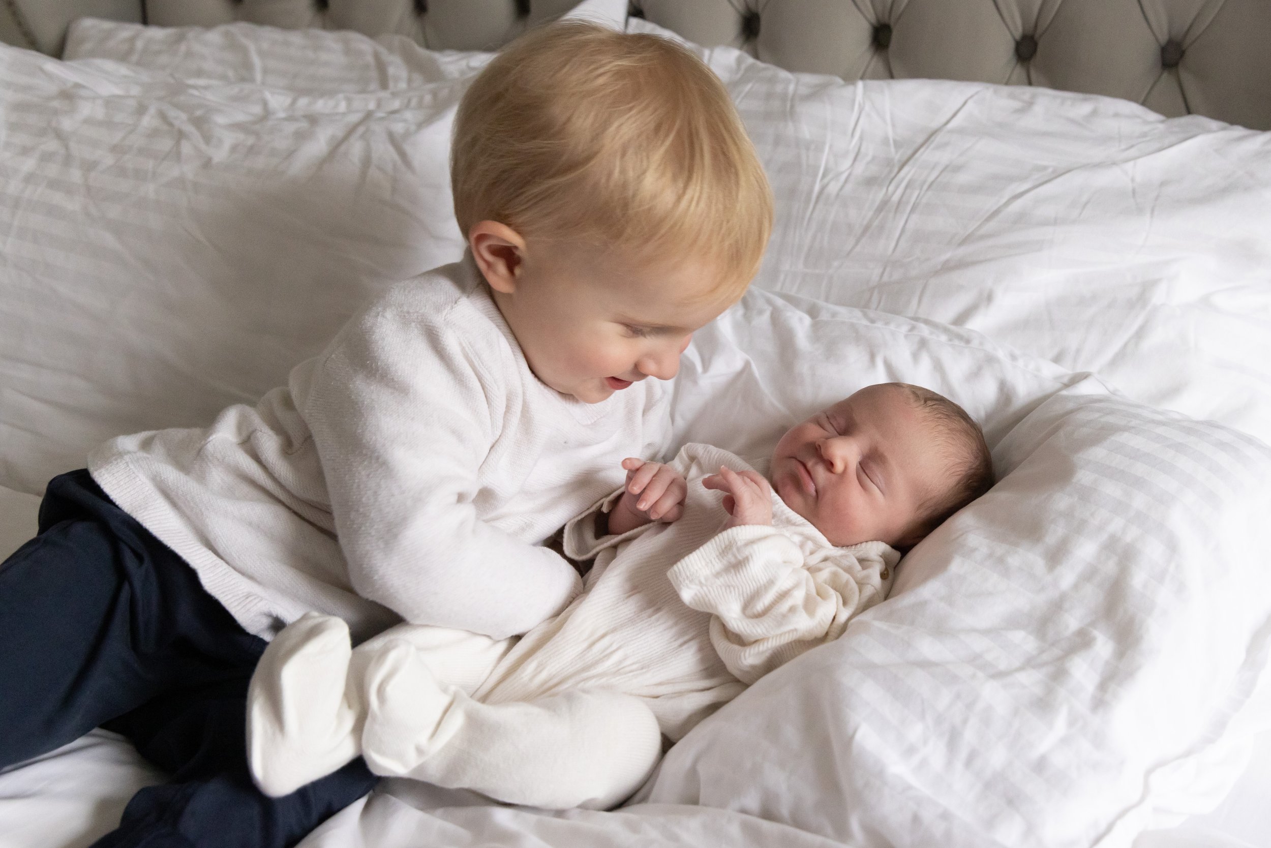 A young boy with blonde hair and a white sweater is lying on a bed with white sheets, holding and looking fondly at a newborn baby who is also lying on the bed. The background features a plush headboard.
