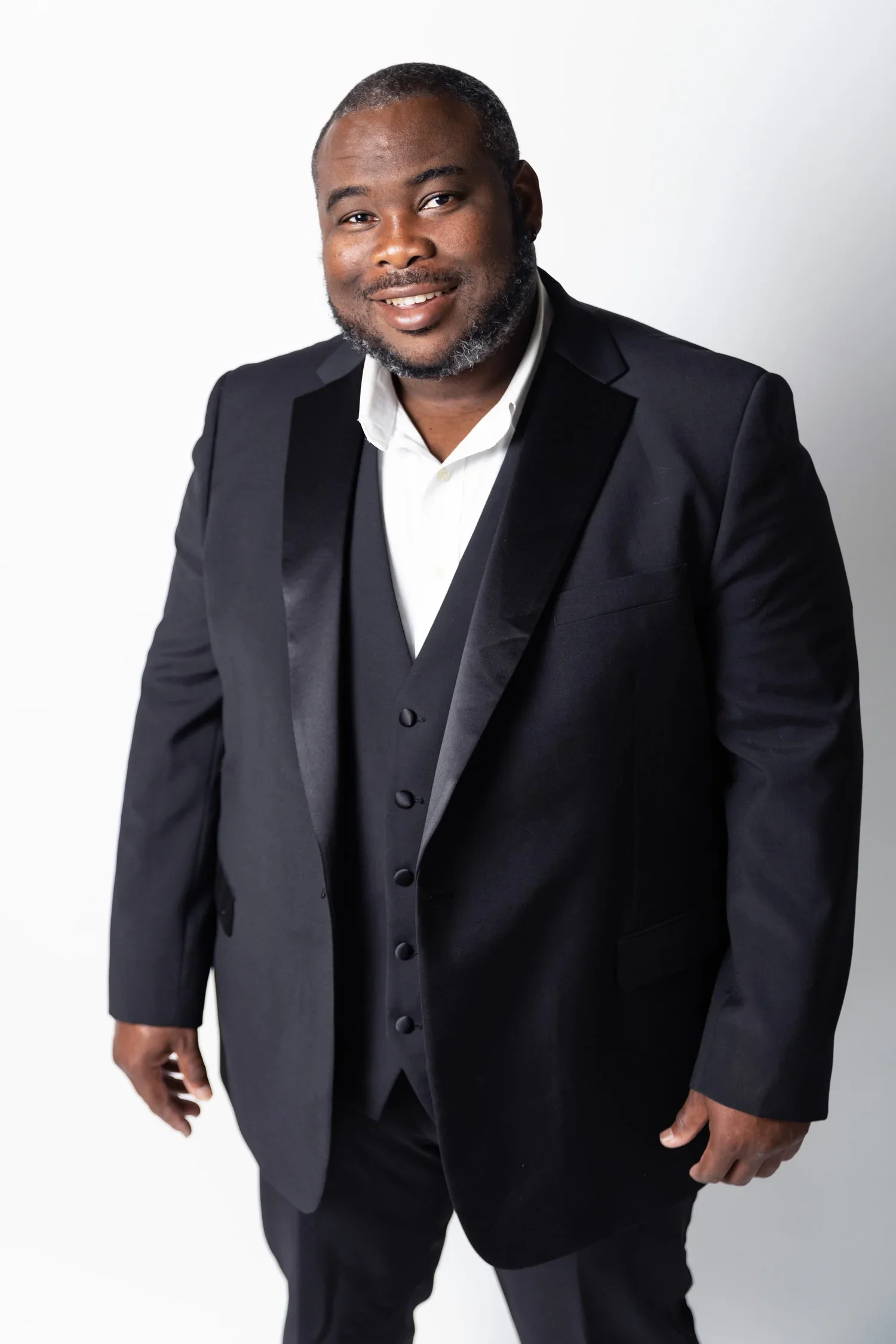 A man in a black tuxedo with a white dress shirt, smiling at the camera against a plain white background.