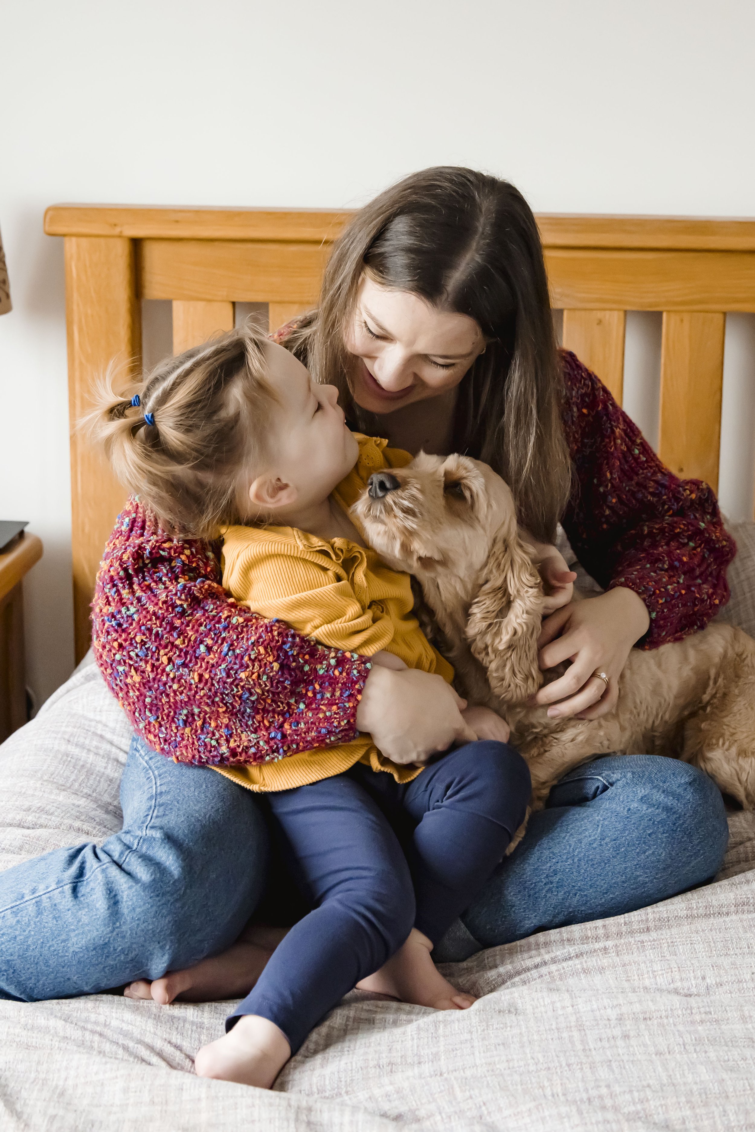A woman and a young girl sitting on a bed, smiling and gazing at each other while holding a small, fluffy dog between them. The girl is wearing a yellow top and blue pants, and the woman is wearing a multicolored sweater.