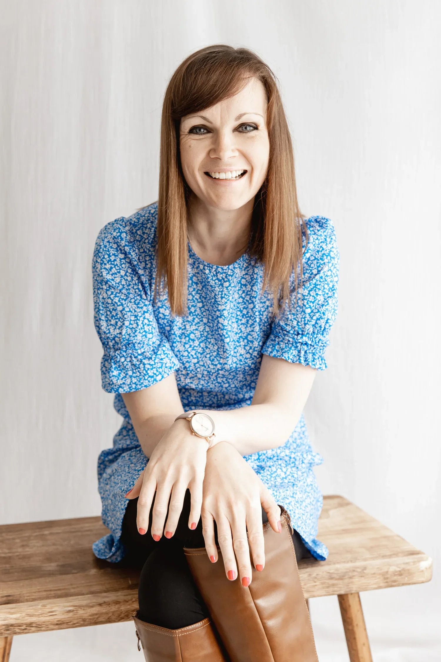 A smiling woman with long brown hair and blue eyes, wearing a blue floral dress, sitting on a wooden bench against a light-colored background.