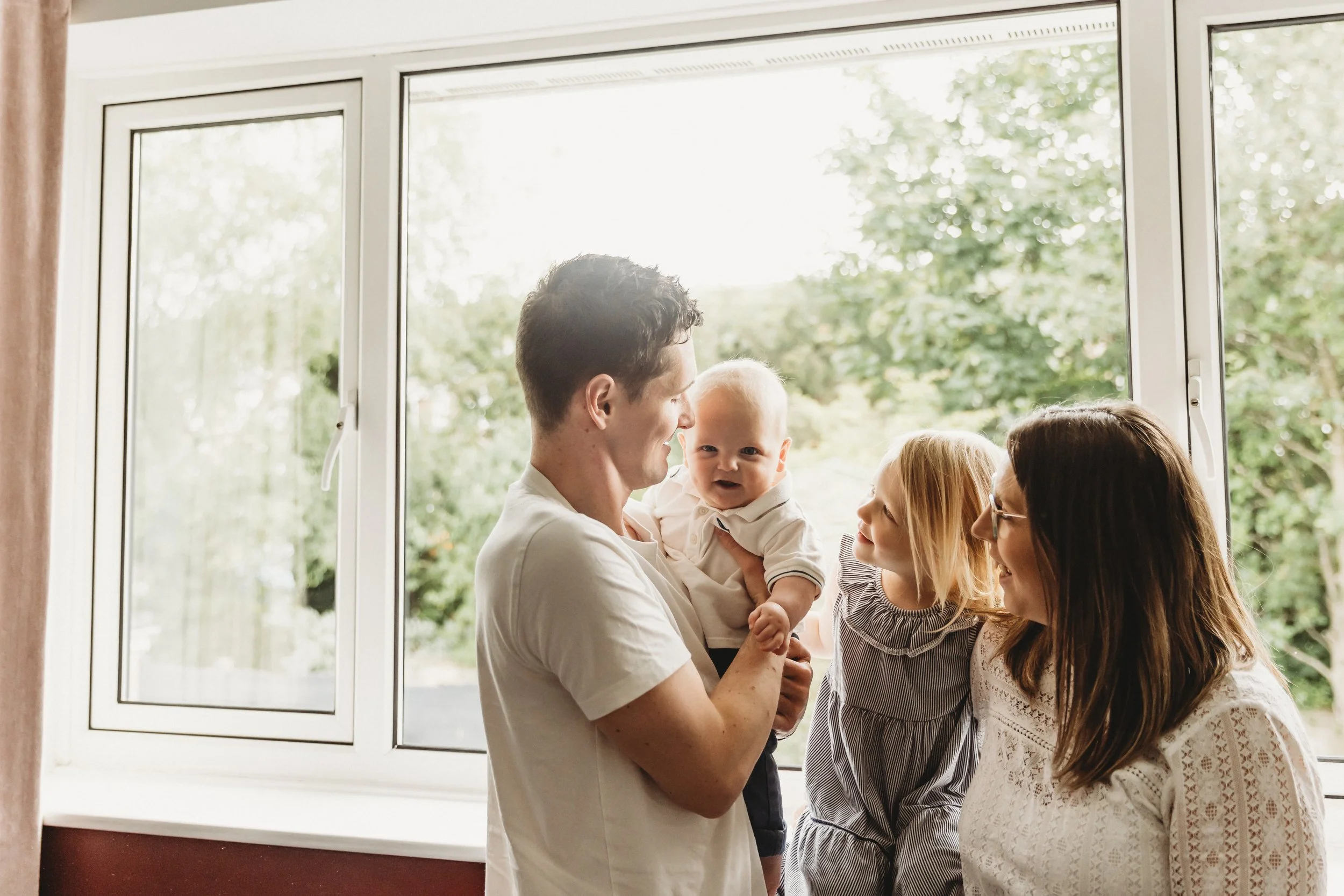 A man holding a baby girl while three women stand nearby, looking at the baby and smiling inside a house near a large window with a view of green trees outside.