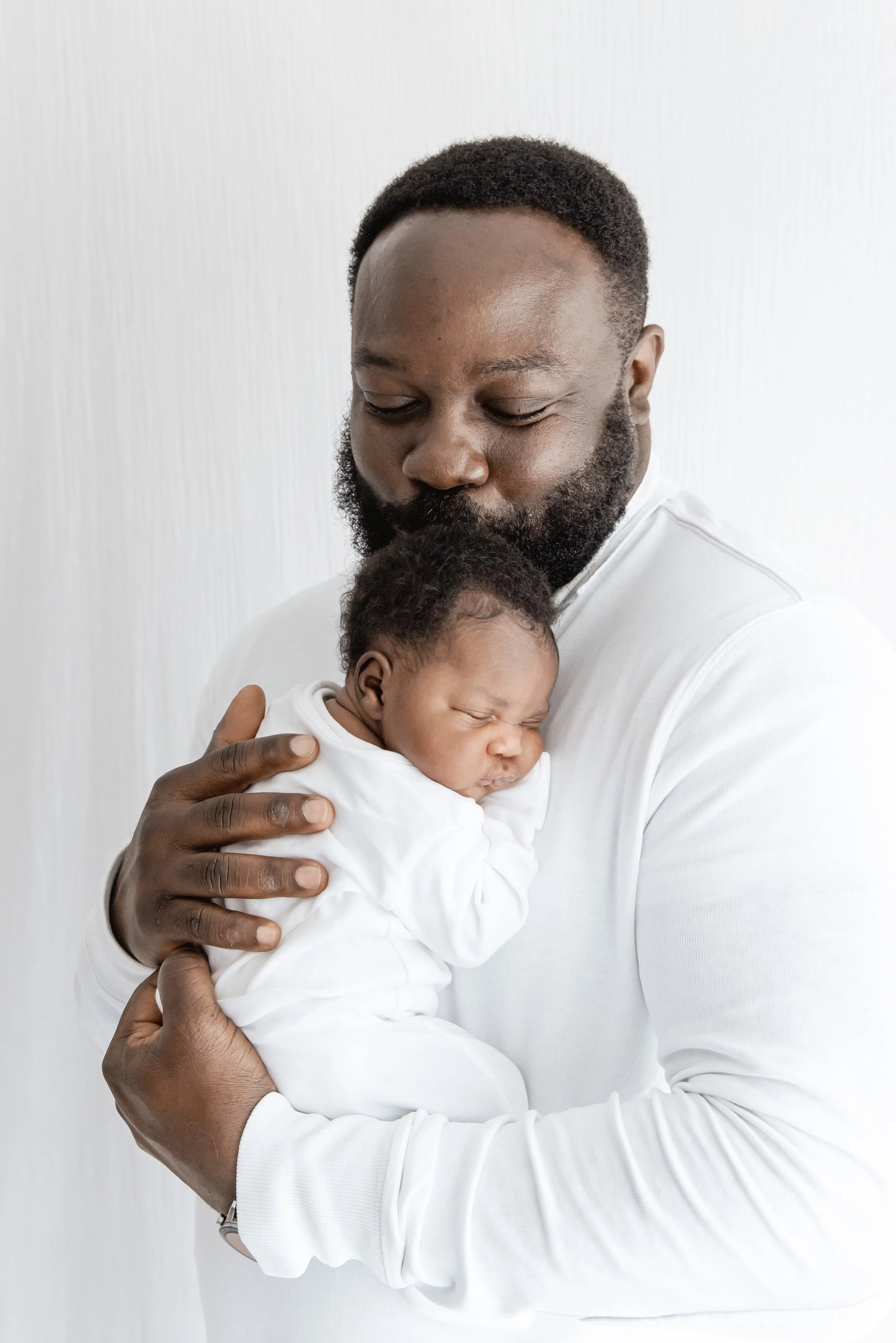A man with a beard and short curly hair holding a sleeping infant child against a plain white wall.