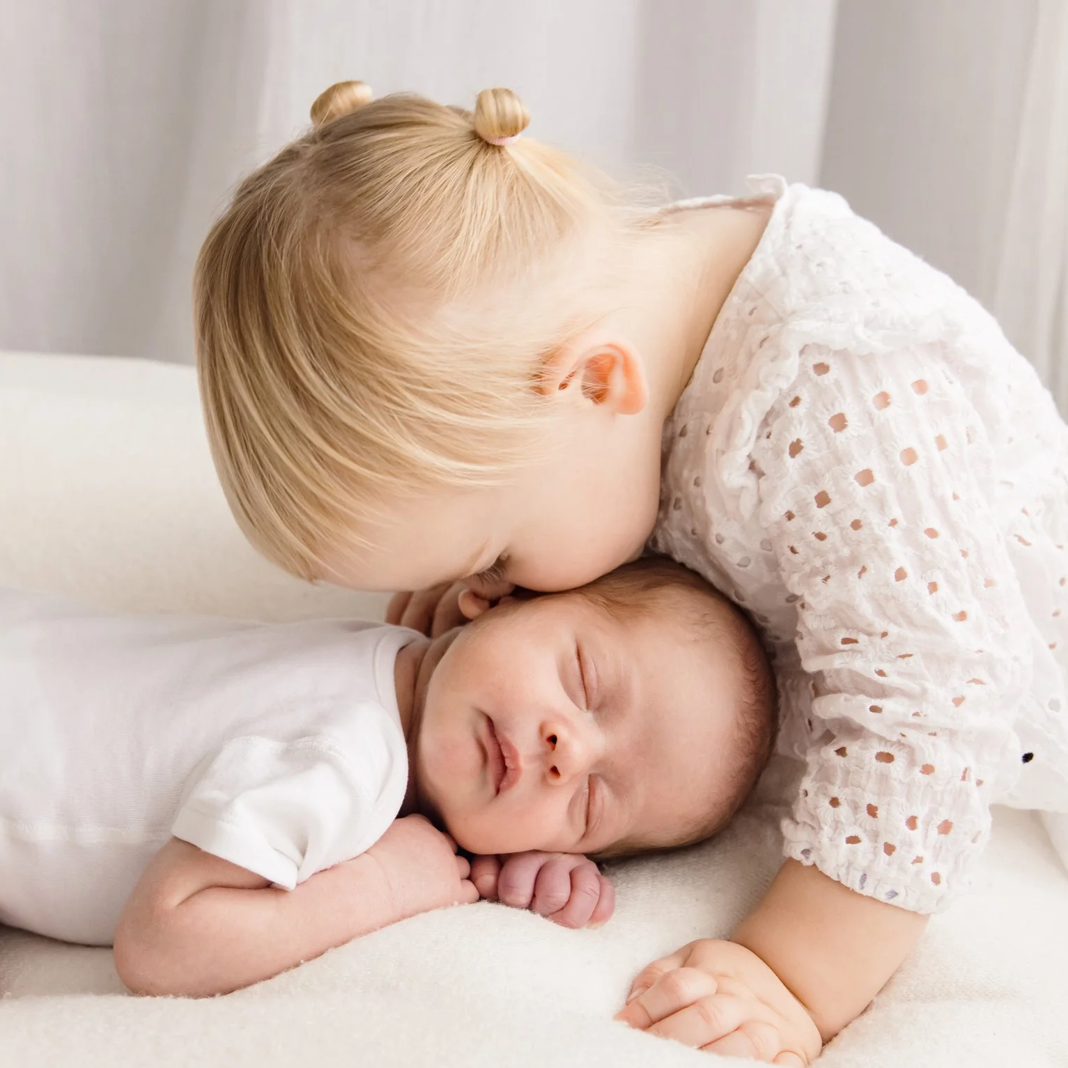A young girl with blonde hair styled in buns kisses a sleeping baby on the forehead. The baby is lying on a soft, white surface.