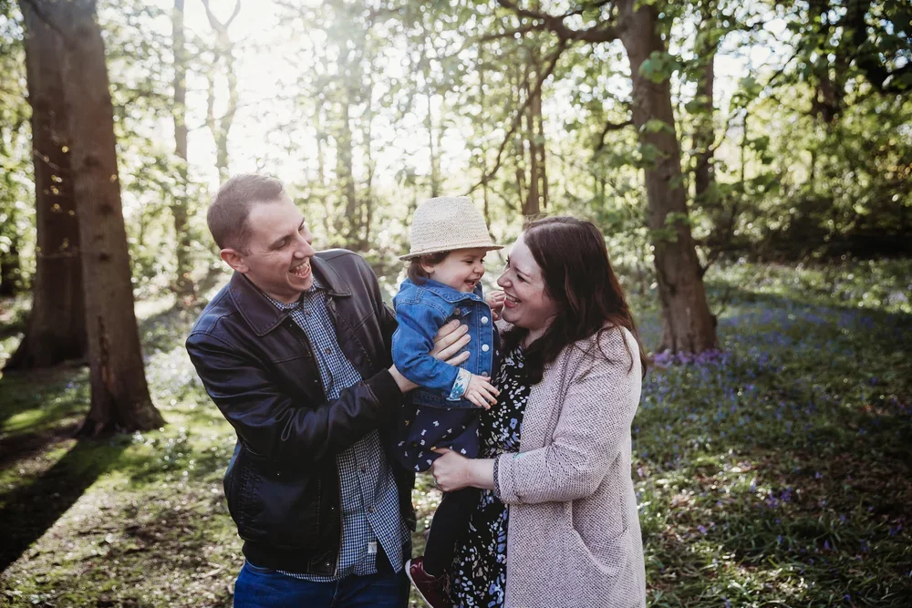 A happy family of three enjoying time outdoors in a forest, with sunlight filtering through trees. The father, mother, and young child are smiling and laughing together.