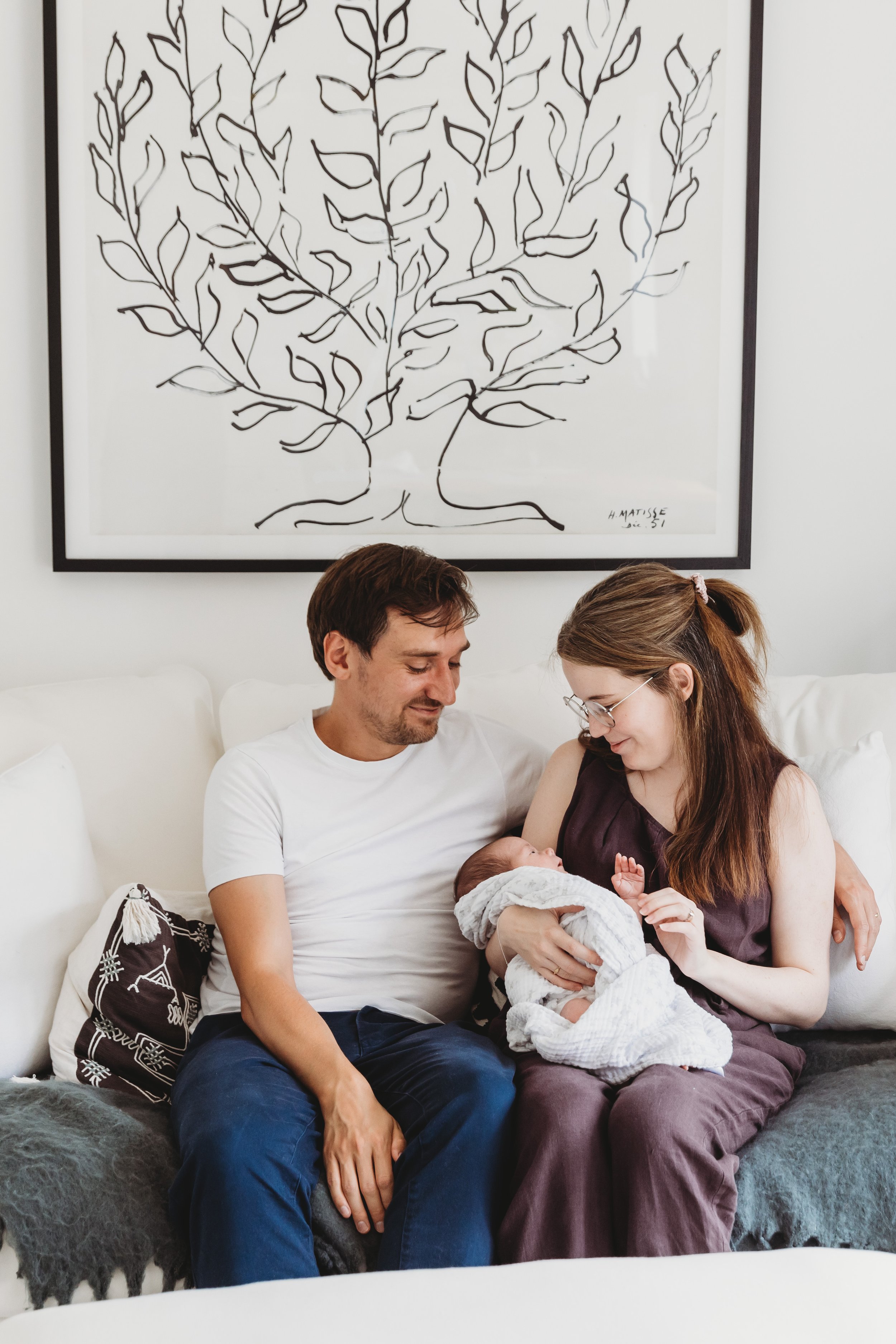 A family of three sitting on a couch breastfeeding their newborn baby. The father has dark hair and a beard, wearing a white t-shirt. The mother has long brown hair, glasses, and is wearing a dark outfit. The baby is wrapped in a white blanket. The b
