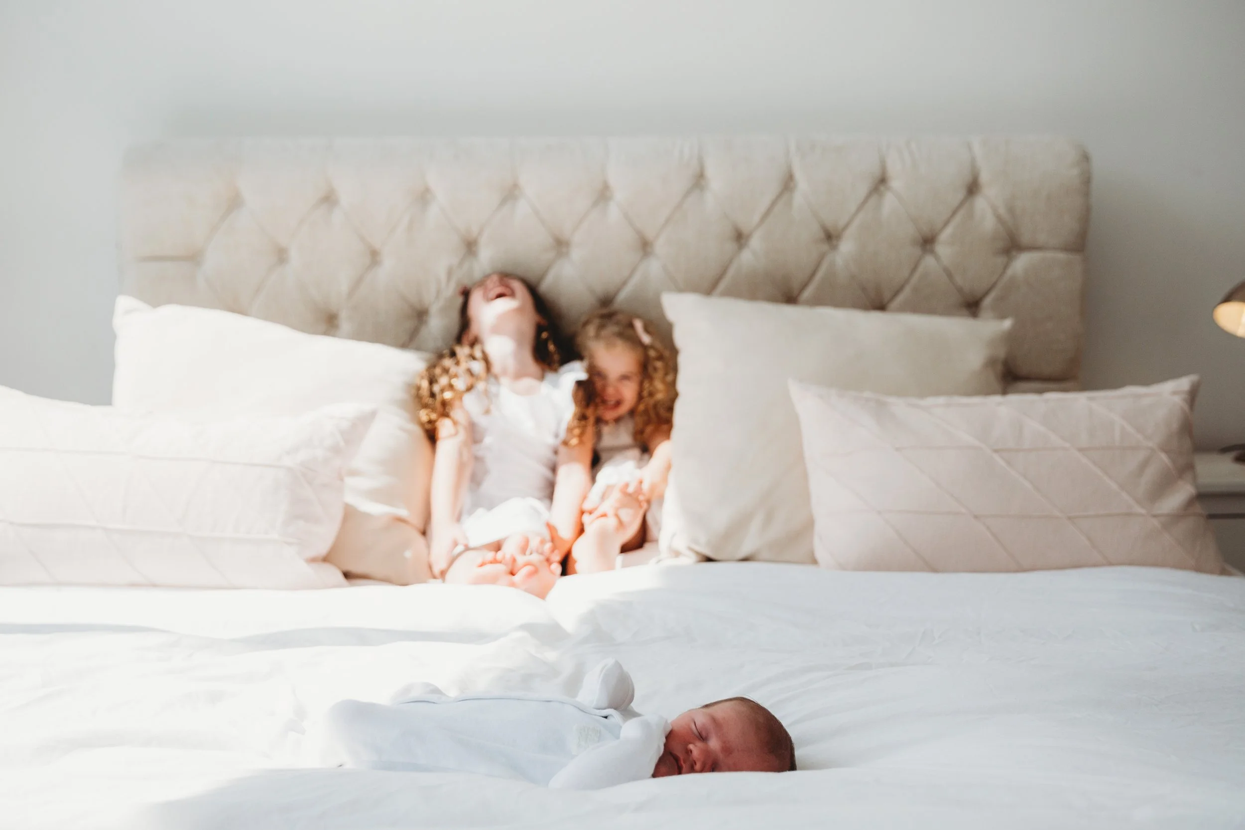Two children with curly hair sitting on a large bed with white bedding and pillows, laughing and playing, with a beige tufted headboard against a plain wall.