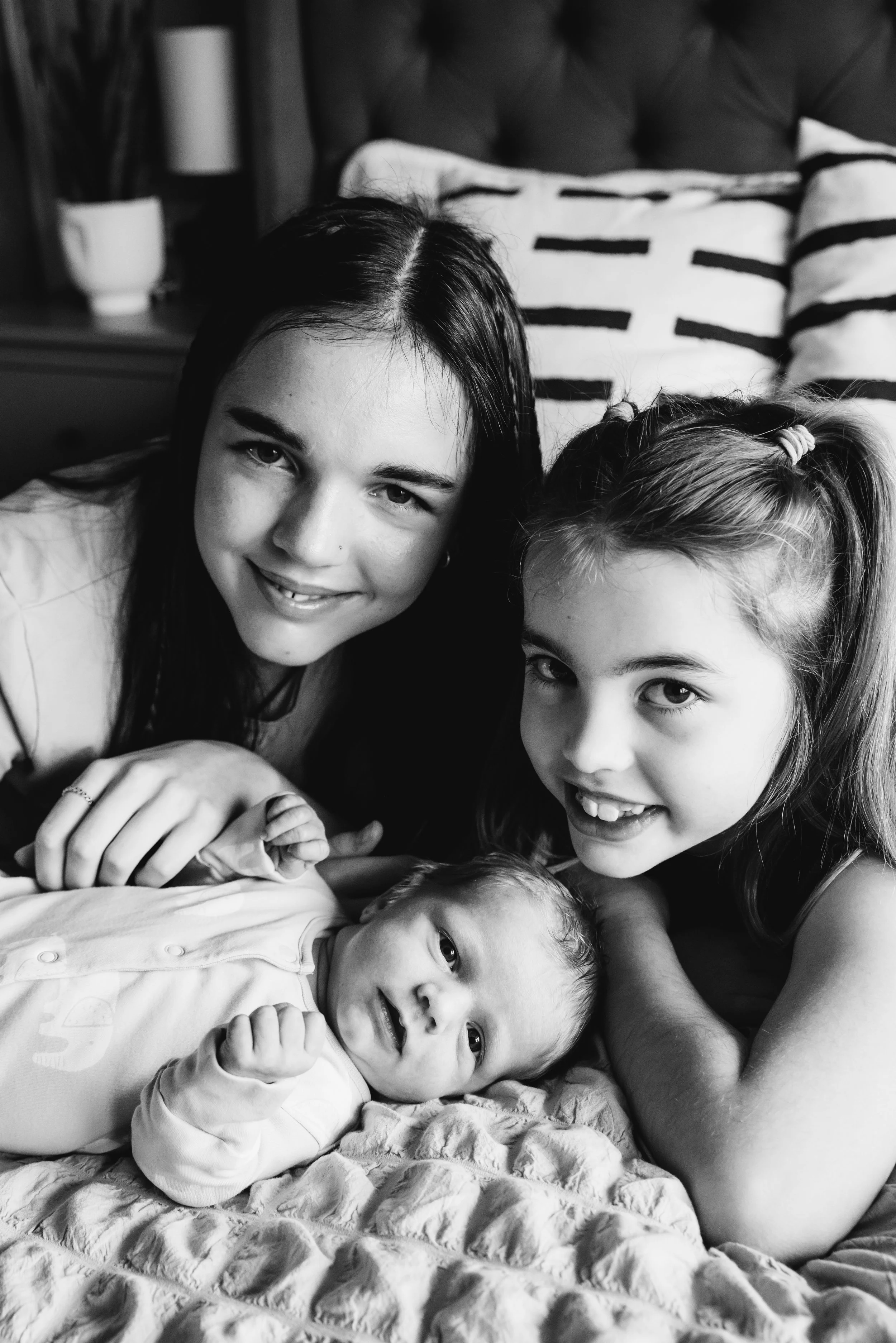 A black and white photo of three females and a baby lying on a bed. The females are smiling at the camera, with two of them having long dark hair and the baby has a light hairstyle. The baby is lying on a quilted blanket in front of the females.