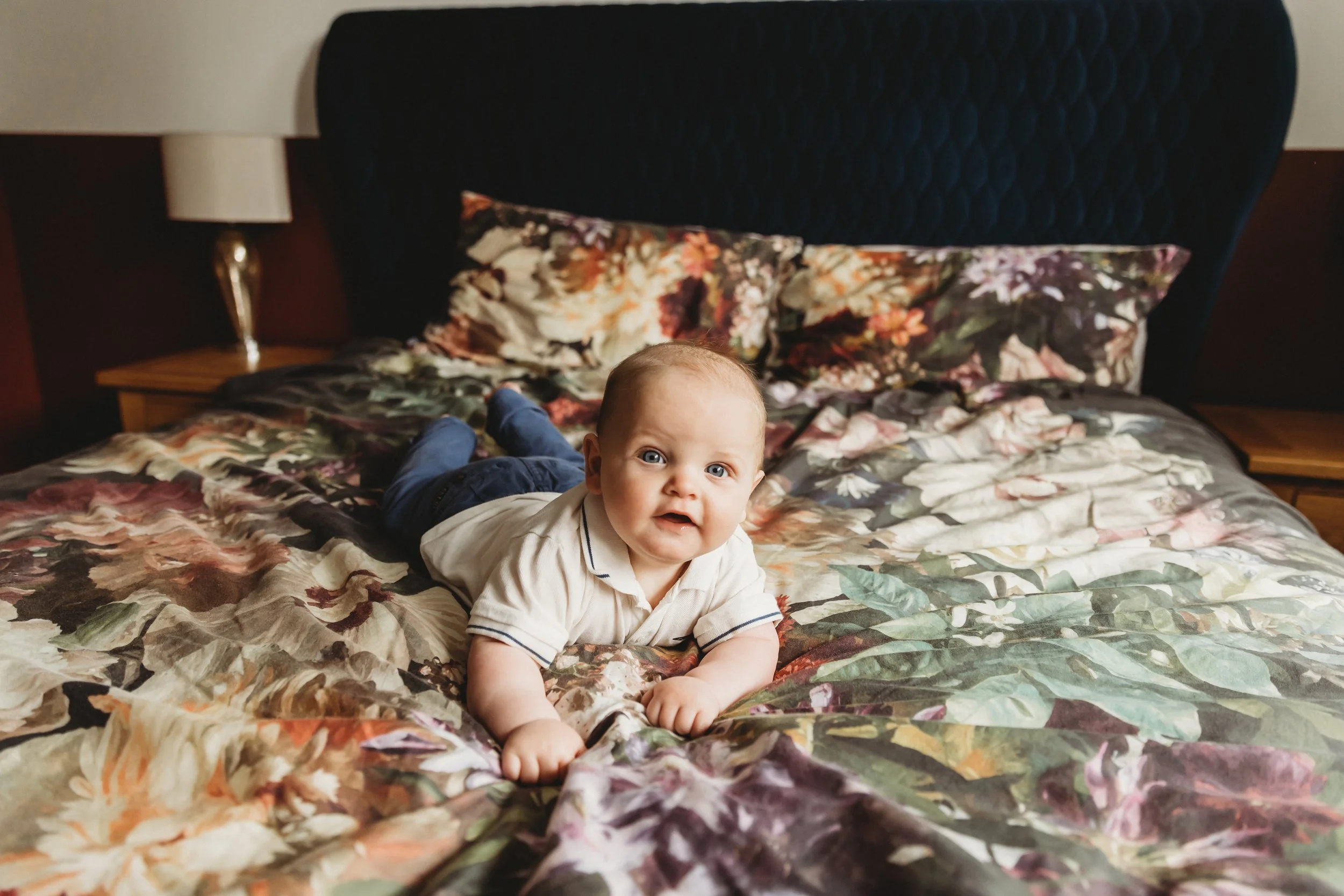 A baby lying on a floral-patterned bedspread, looking at the camera with a curious expression.