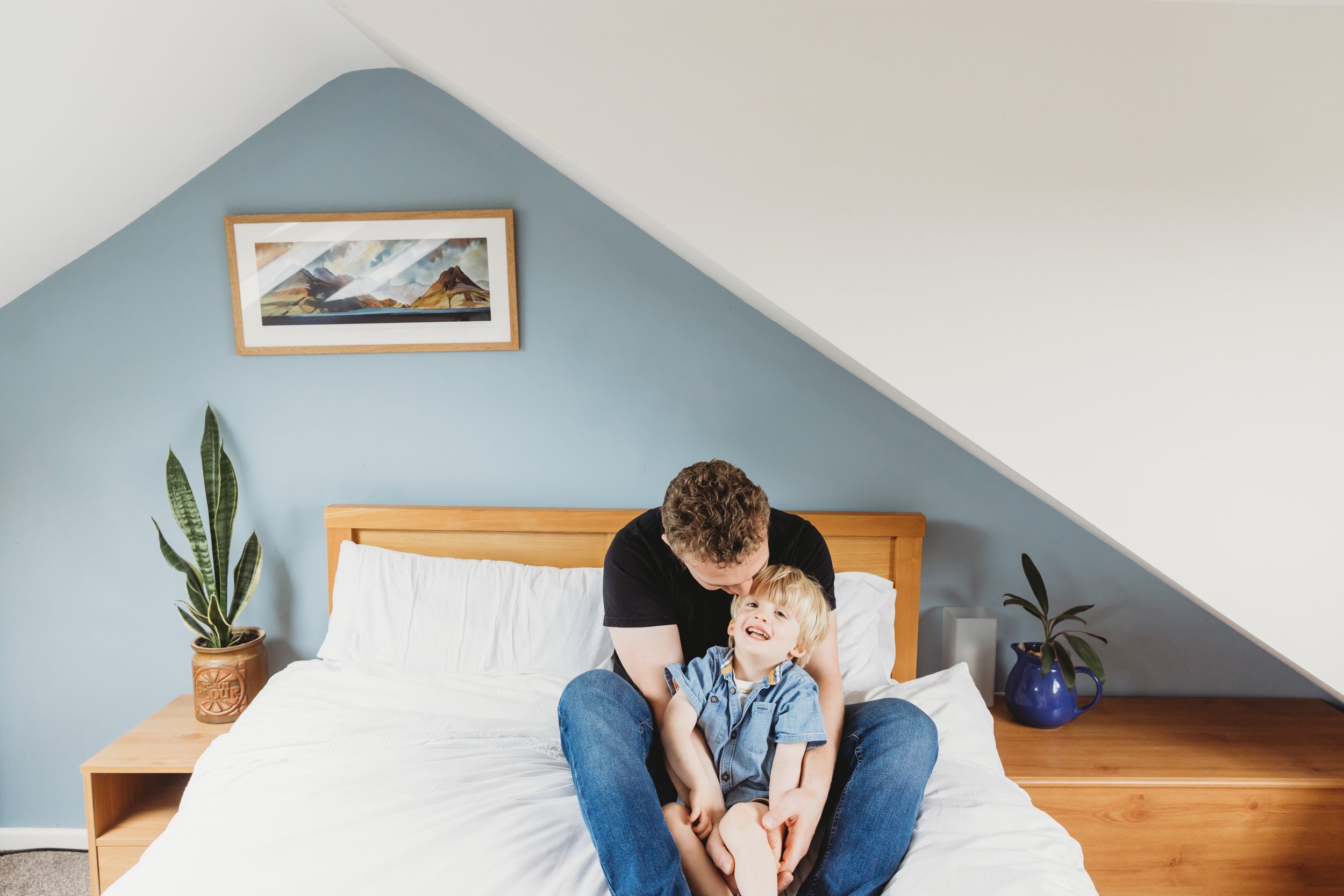 A man and a young boy sitting on a bed in a bedroom with a sloped ceiling. The man is leaning forward with his head close to the boy, both are smiling and wearing casual clothes. There are two potted plants on either side of the bed, one in a clay po