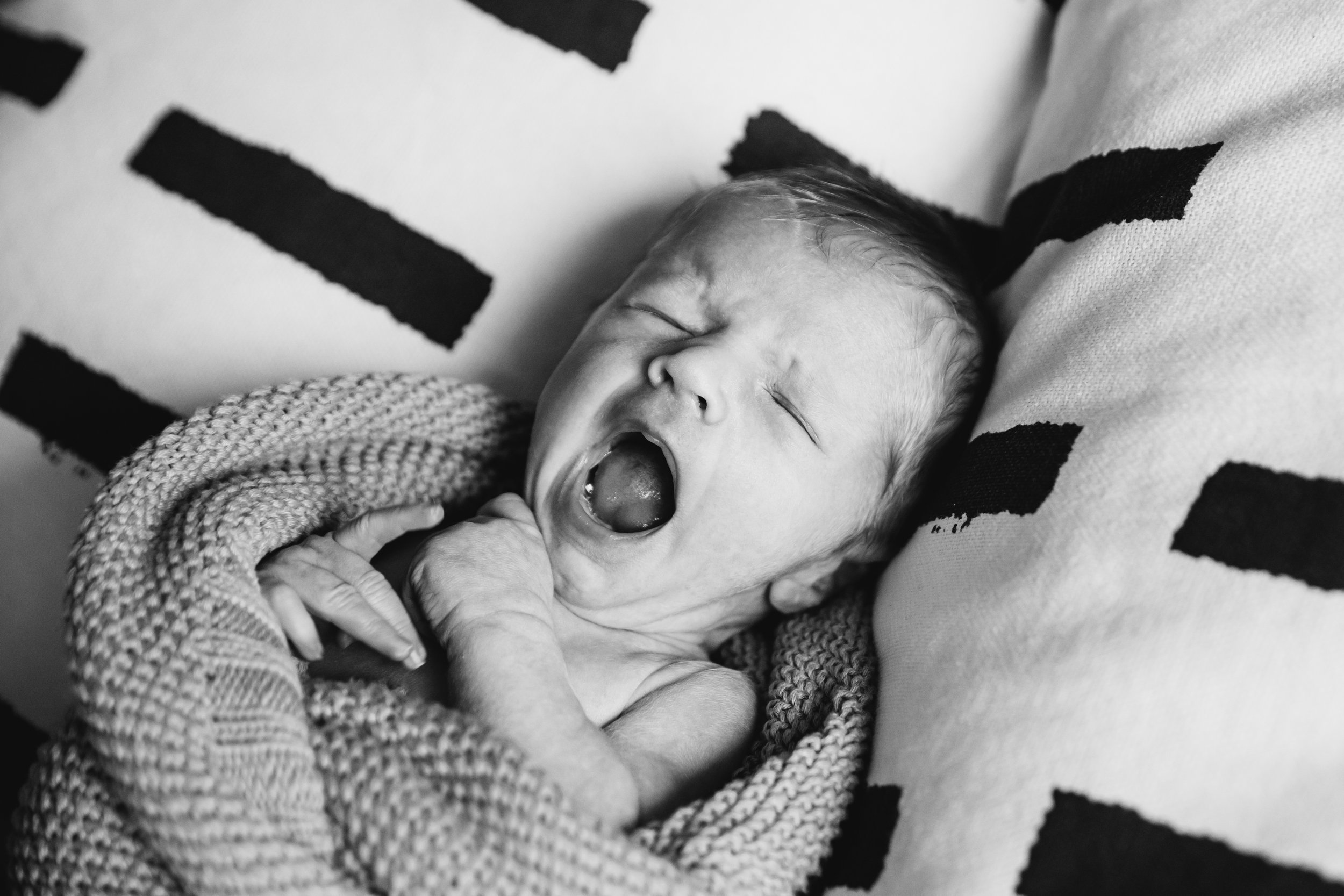 Black and white photo of a young child yawning, wearing a chunky knit sweater, lying on a patterned pillow with bold black stripes.