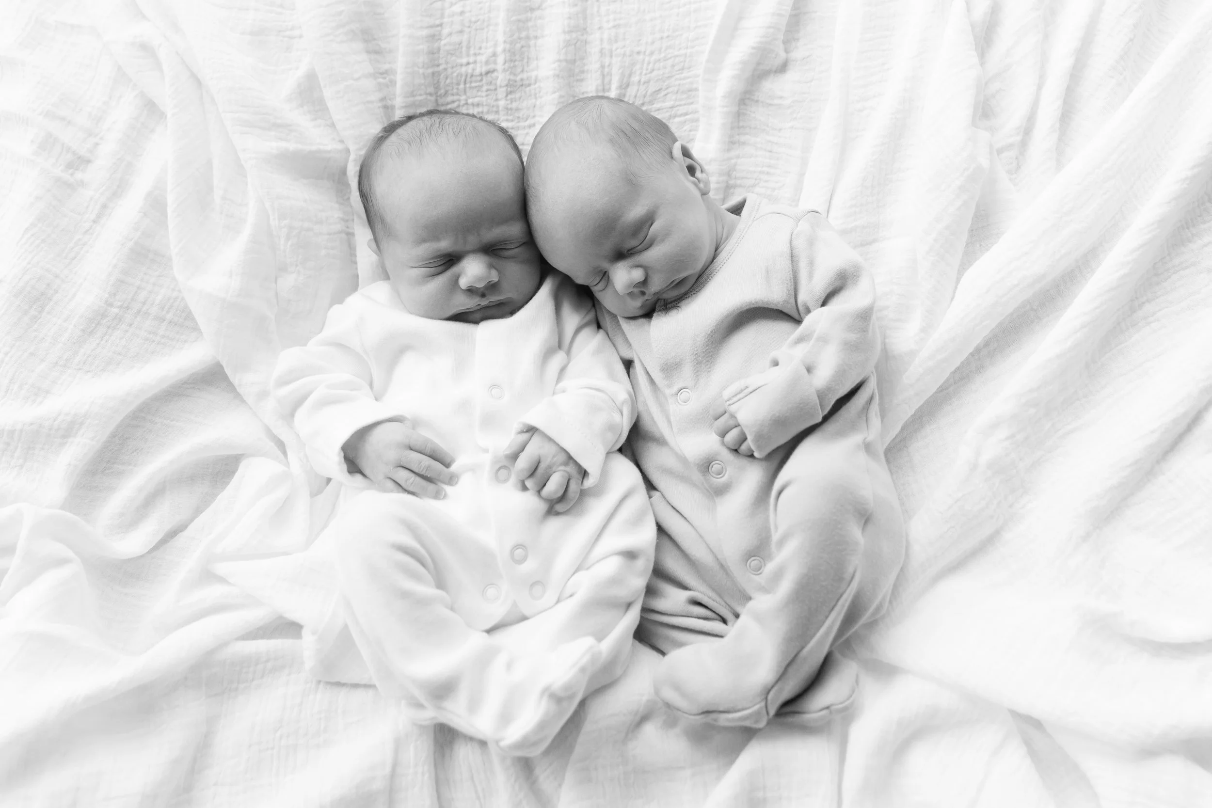Two sleeping infants lying close together on a bed with white sheets, curled up and touching heads.