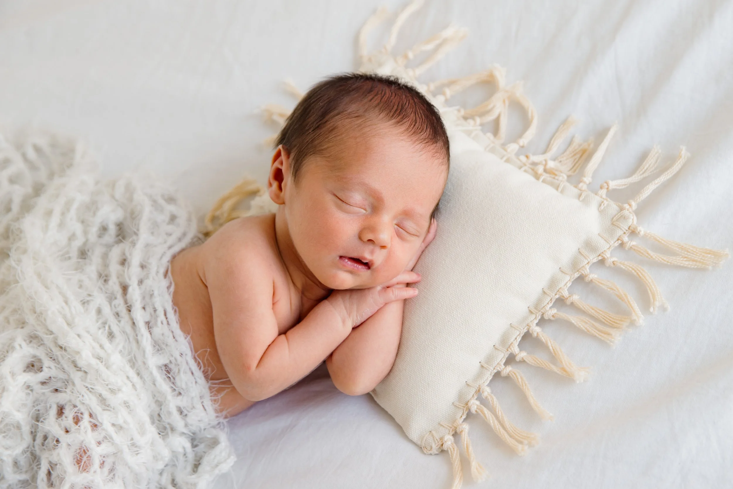A newborn baby with dark hair is peacefully sleeping on a white soft surface, resting its head on a cream-colored pillow with tassels. The baby is partially covered with a fluffy, cream-colored blanket.