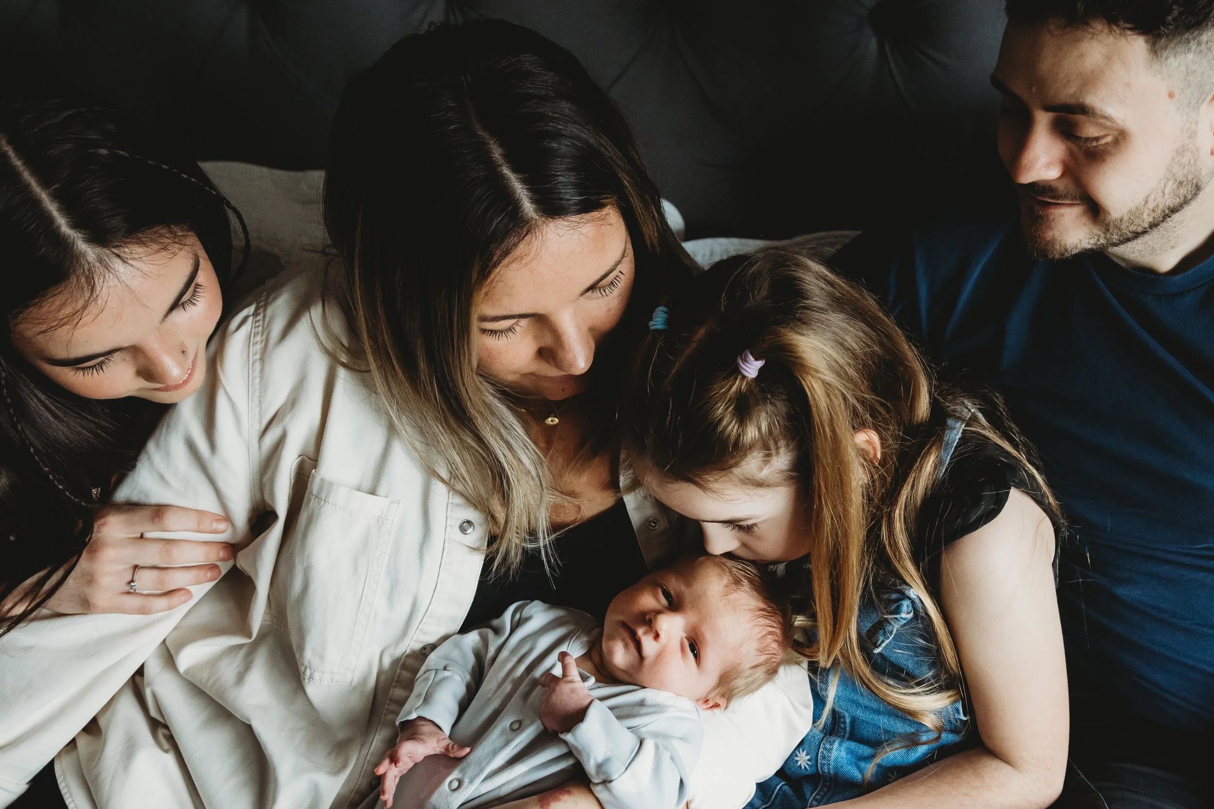 A group of five people, including children and adults, huddled closely together, smiling and looking at each other in a warm, intimate setting.