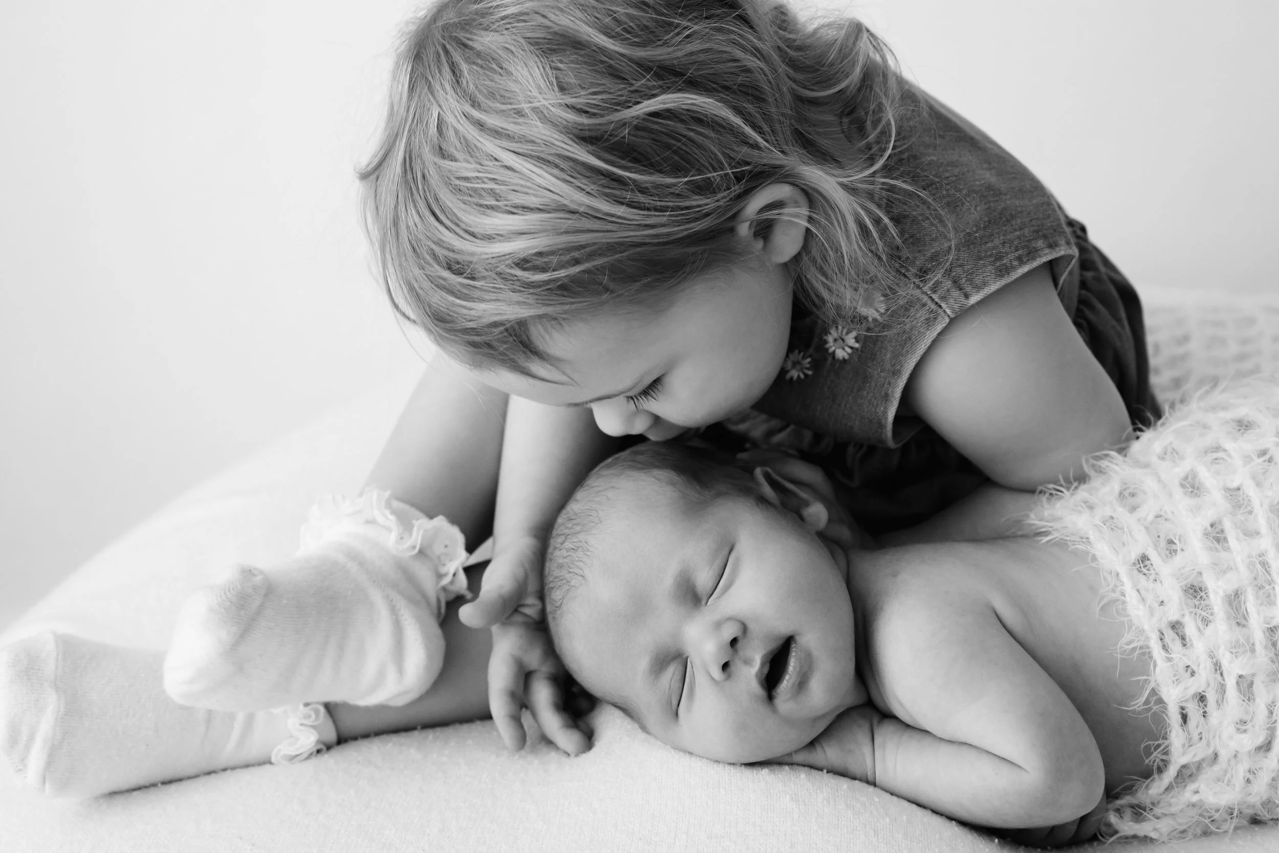 Black and white photograph of a young girl lovingly holding and kissing her sleeping baby sibling, who is lying on a soft surface.