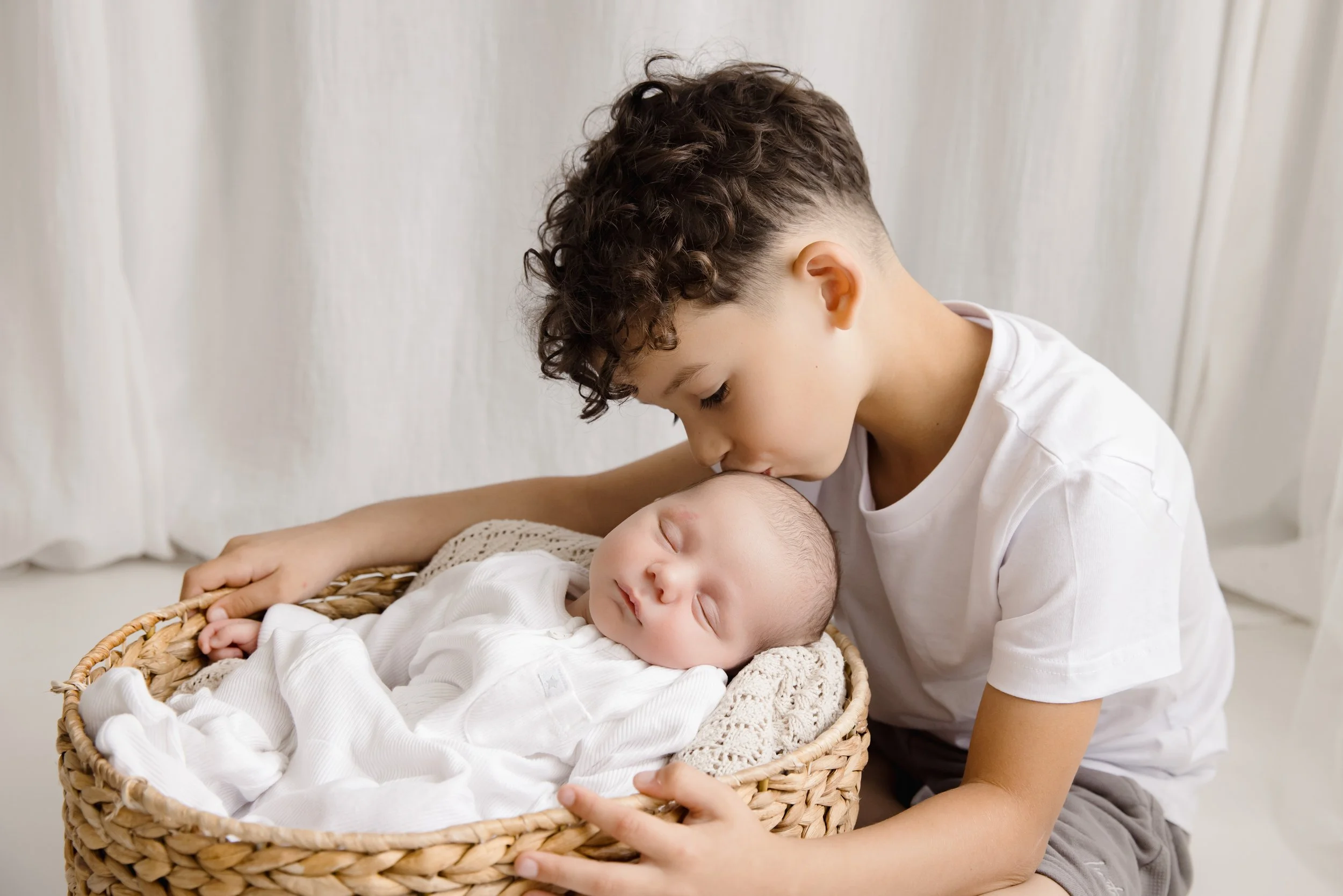 A young boy with short curly hair leaning over and kissing a sleeping baby in a wicker basket.