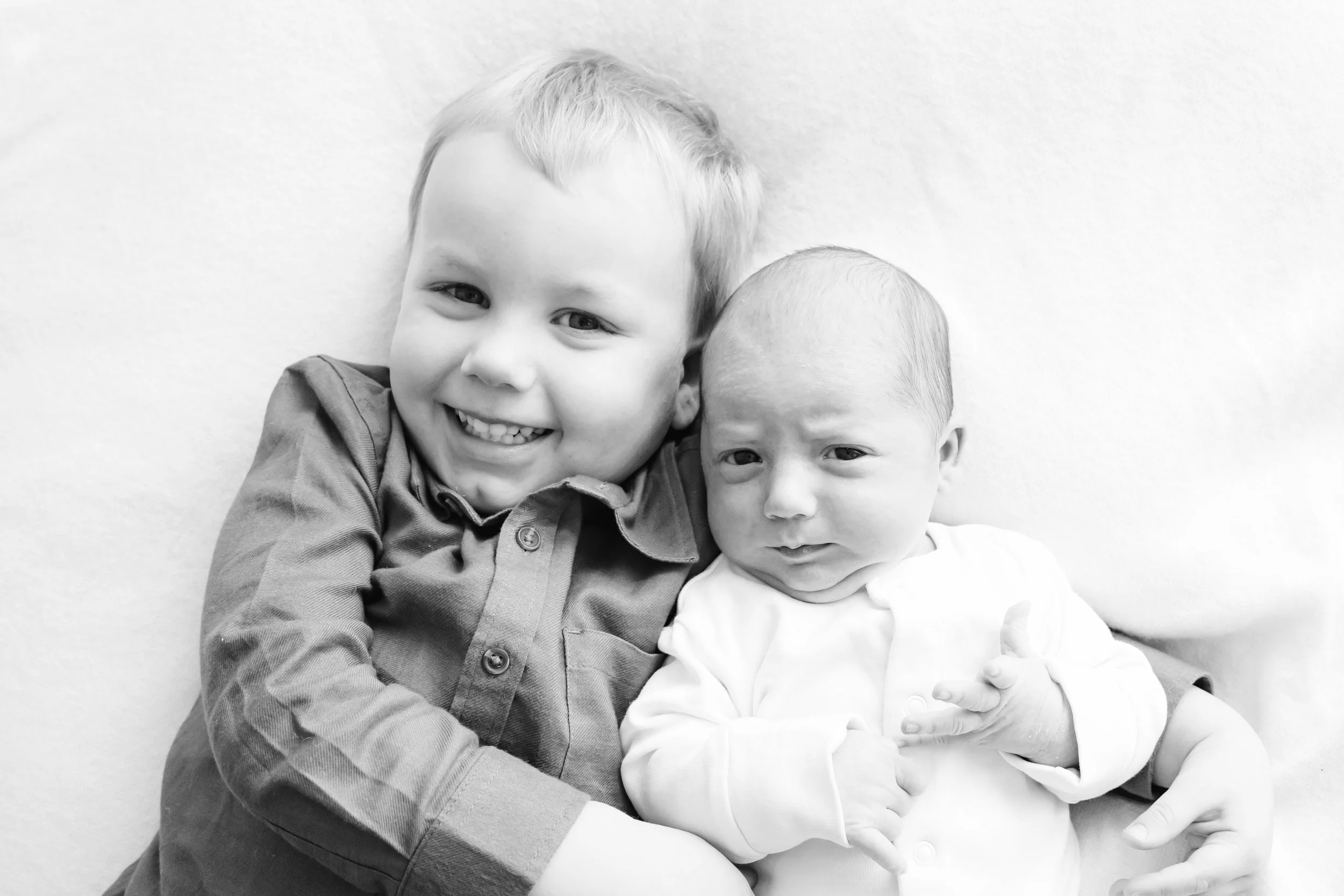 Two young boys, one with light hair and smiling, the other with darker hair and a curious expression, sitting close together against a plain wall.