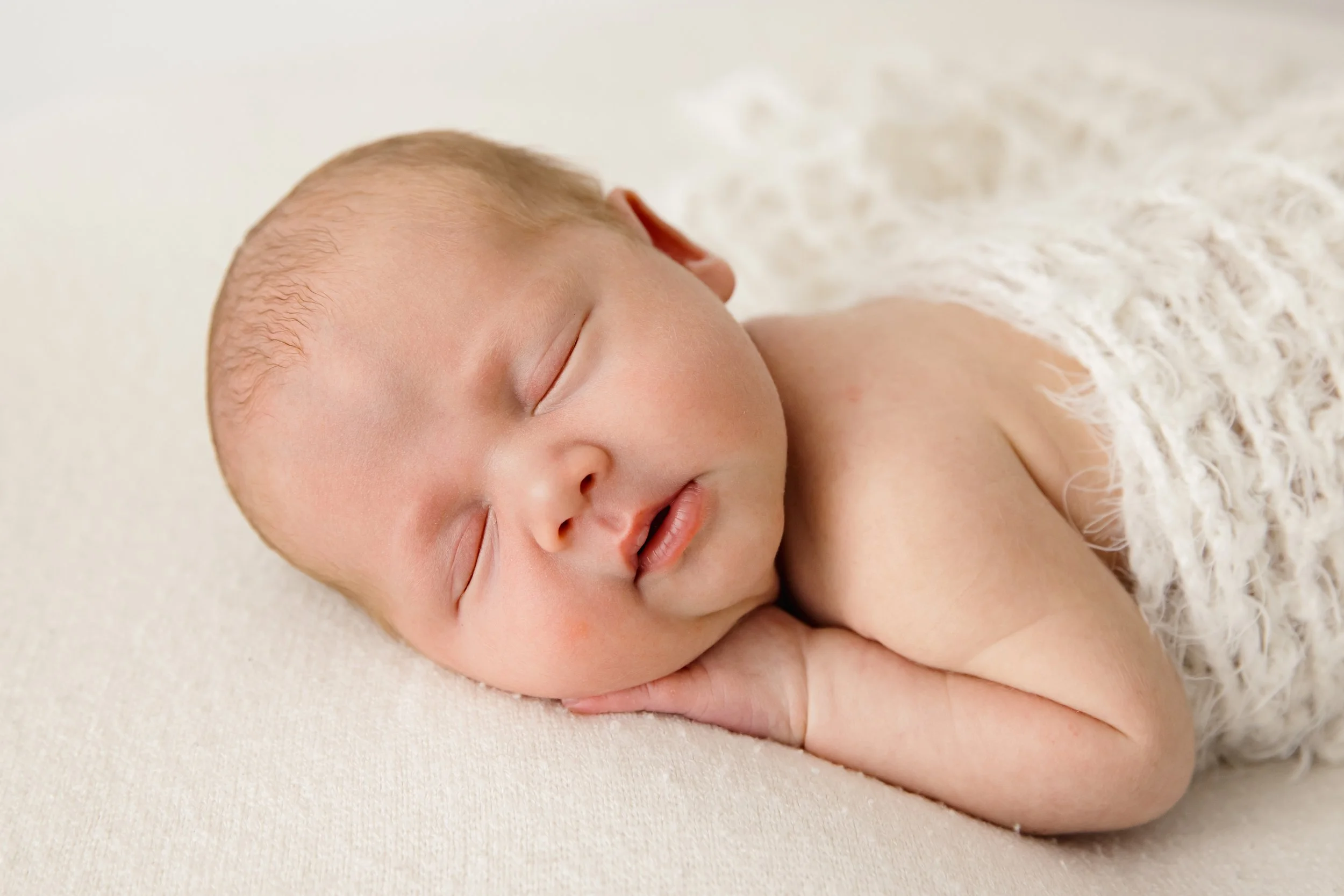 A sleeping baby with light skin, lying on a soft surface, partly covered with a fluffy white blanket.