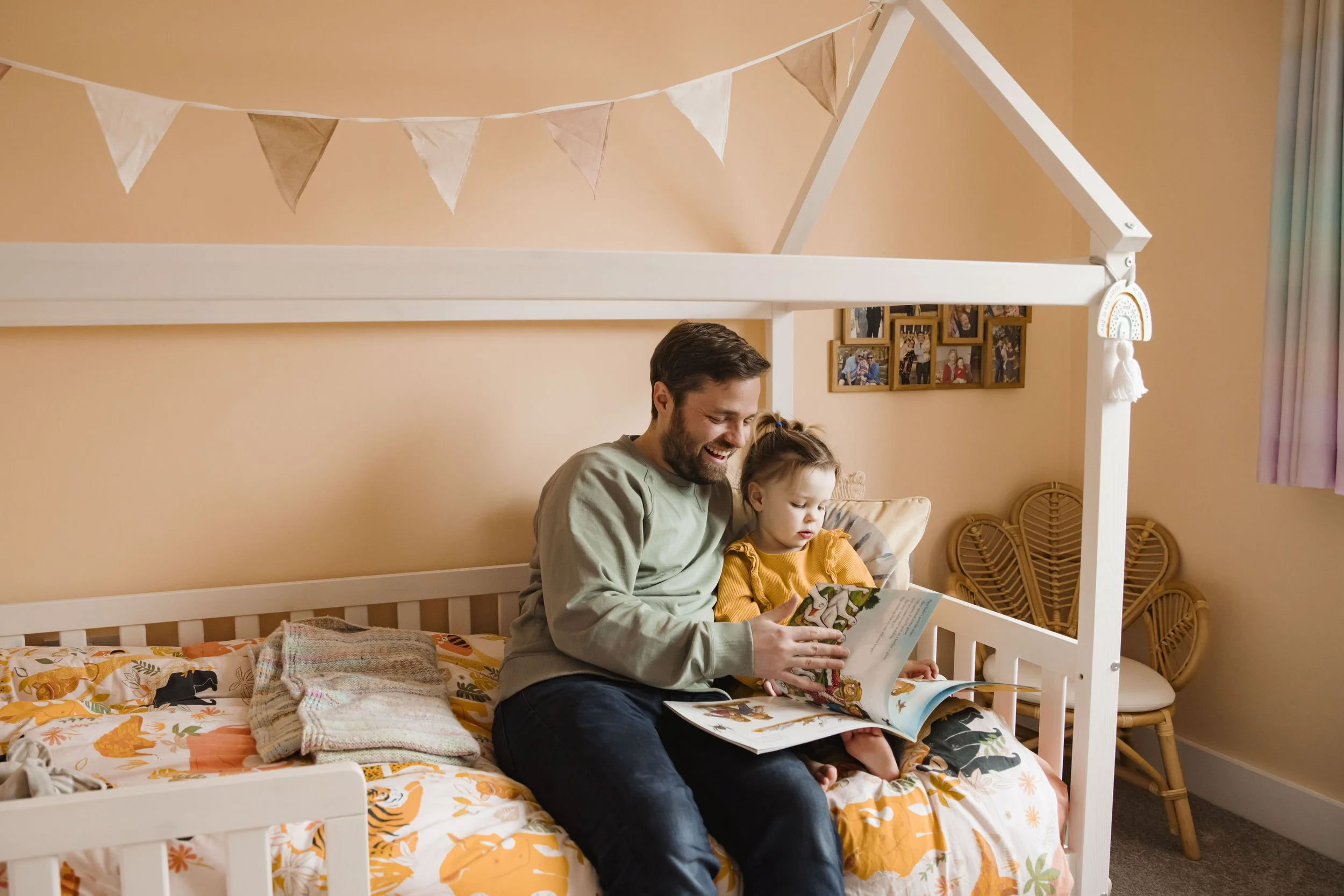 A man and a young girl sitting on a bed in a cozy bedroom, reading a book together. The bed has a colorful, nature-themed bedspread with orange, yellow, and green accents. The walls are painted a soft peach color, and there is a cluster of framed pho
