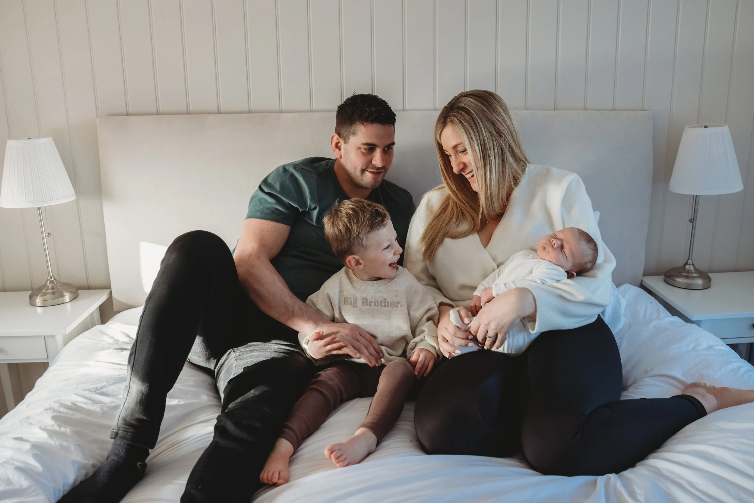 A family of four sitting on a bed, smiling and looking at their newborn baby girl. The parents are sitting close together, with the mother holding the baby and the father leaning in. A young boy is sitting between the parents, smiling at the baby, in