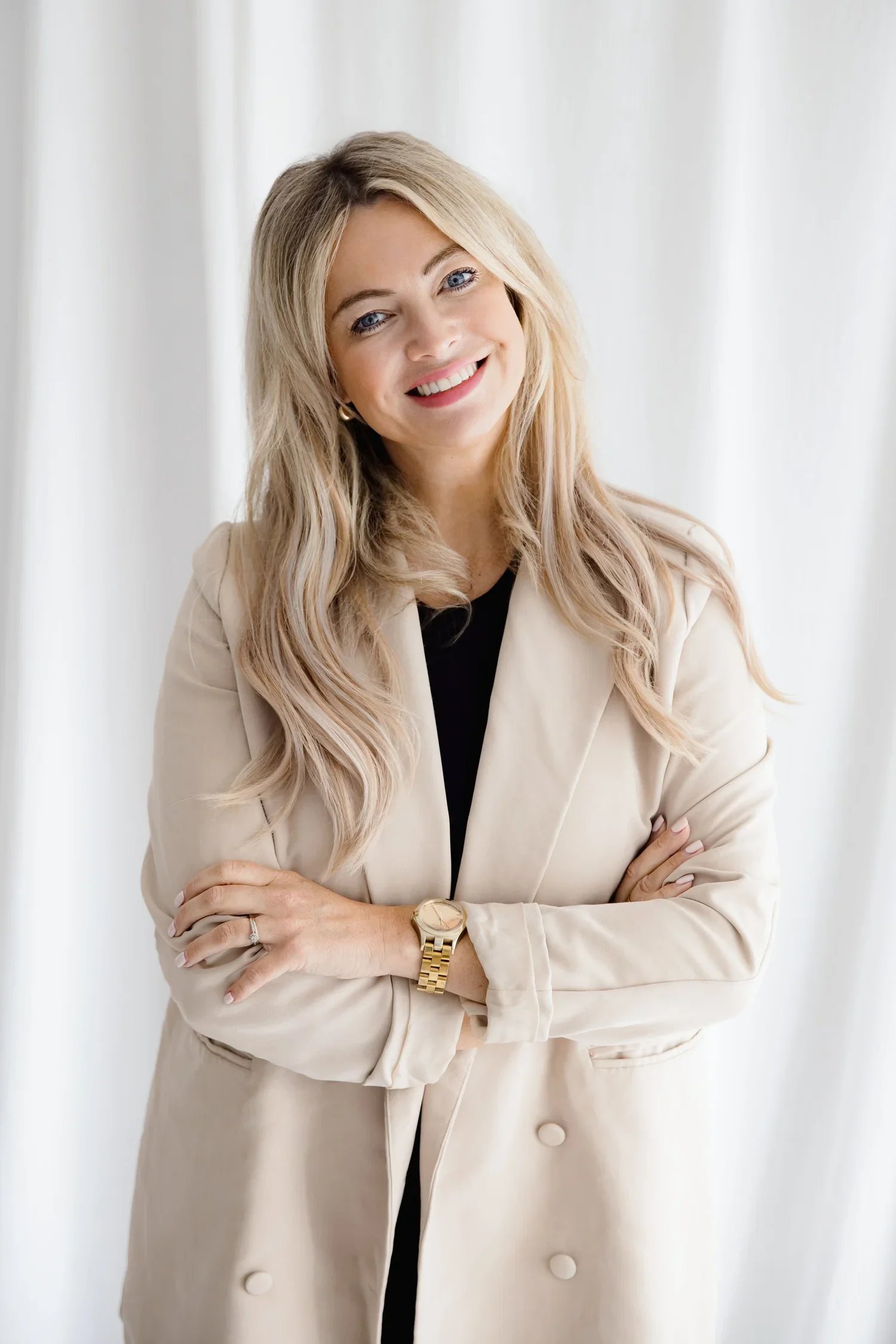 A woman with long blonde hair wearing a beige blazer and a black top, smiling with arms crossed, standing in front of white curtains.