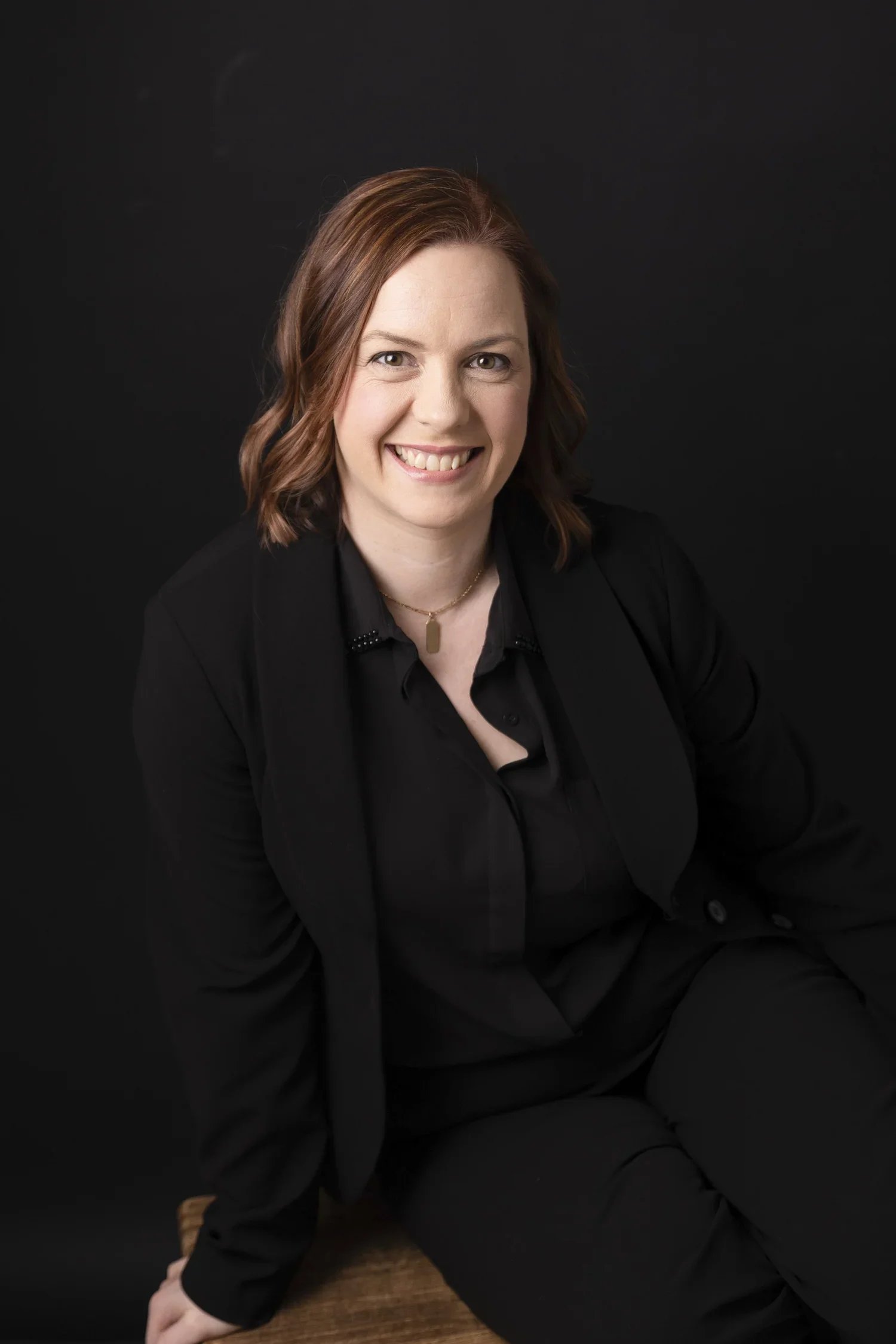 A woman with shoulder-length reddish-brown hair, smiling, dressed in black, sitting on a wooden surface against a dark background.