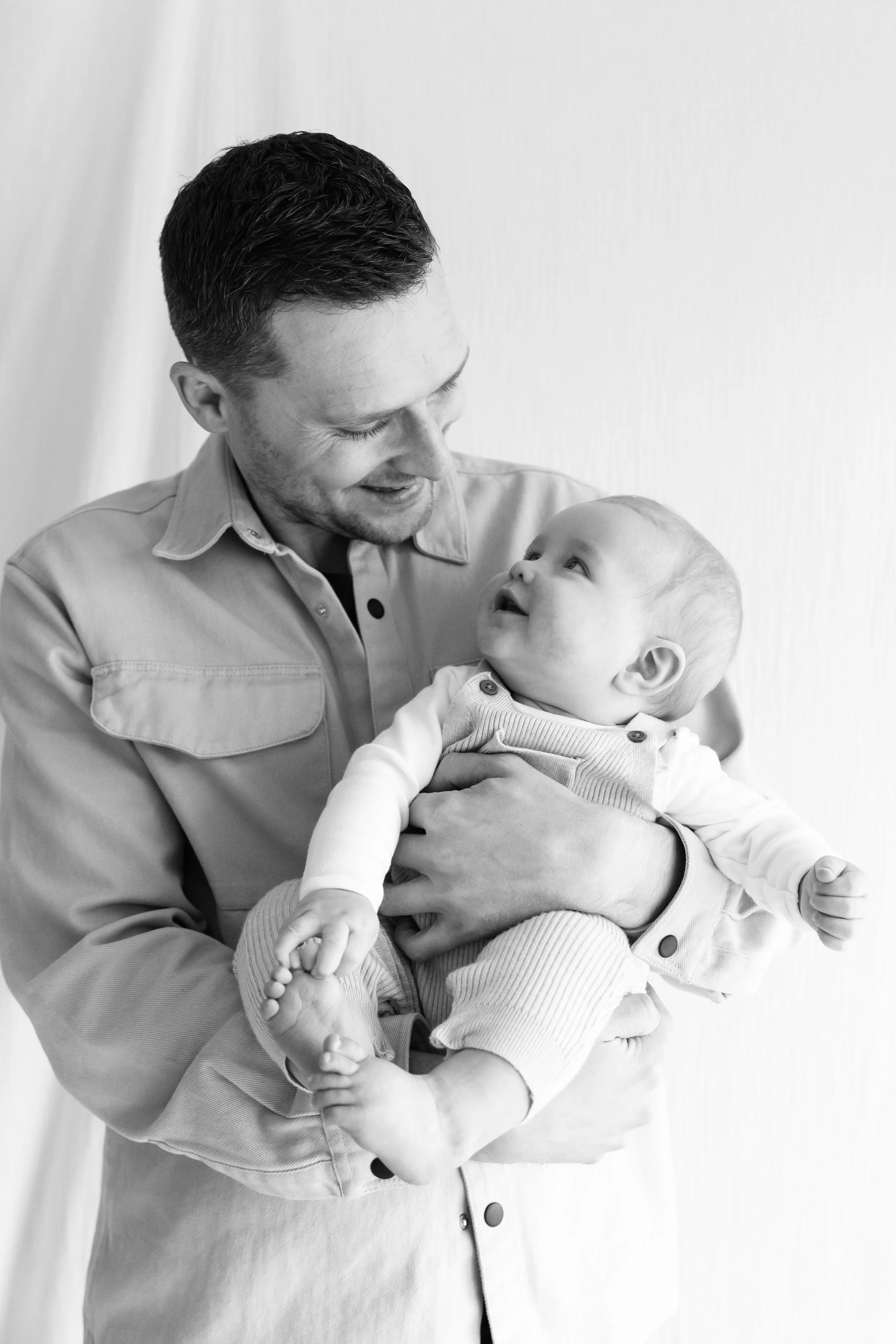 A man smiling and holding a baby in his arms, both looking at each other, in black and white, indoors against a plain background.