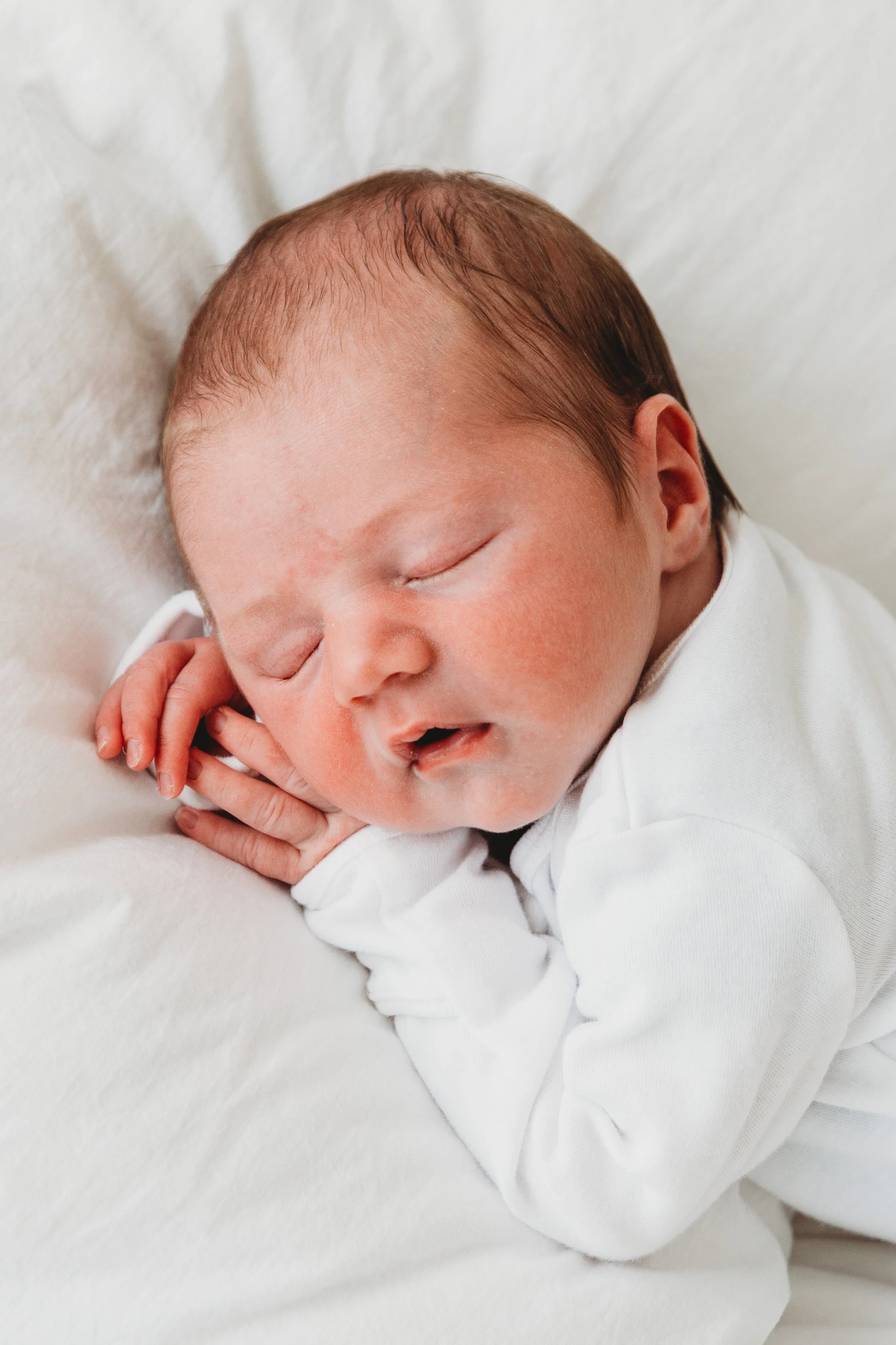 Close-up of a sleeping newborn baby with closed eyes, holding their tiny hand near their face, lying on a soft white surface.