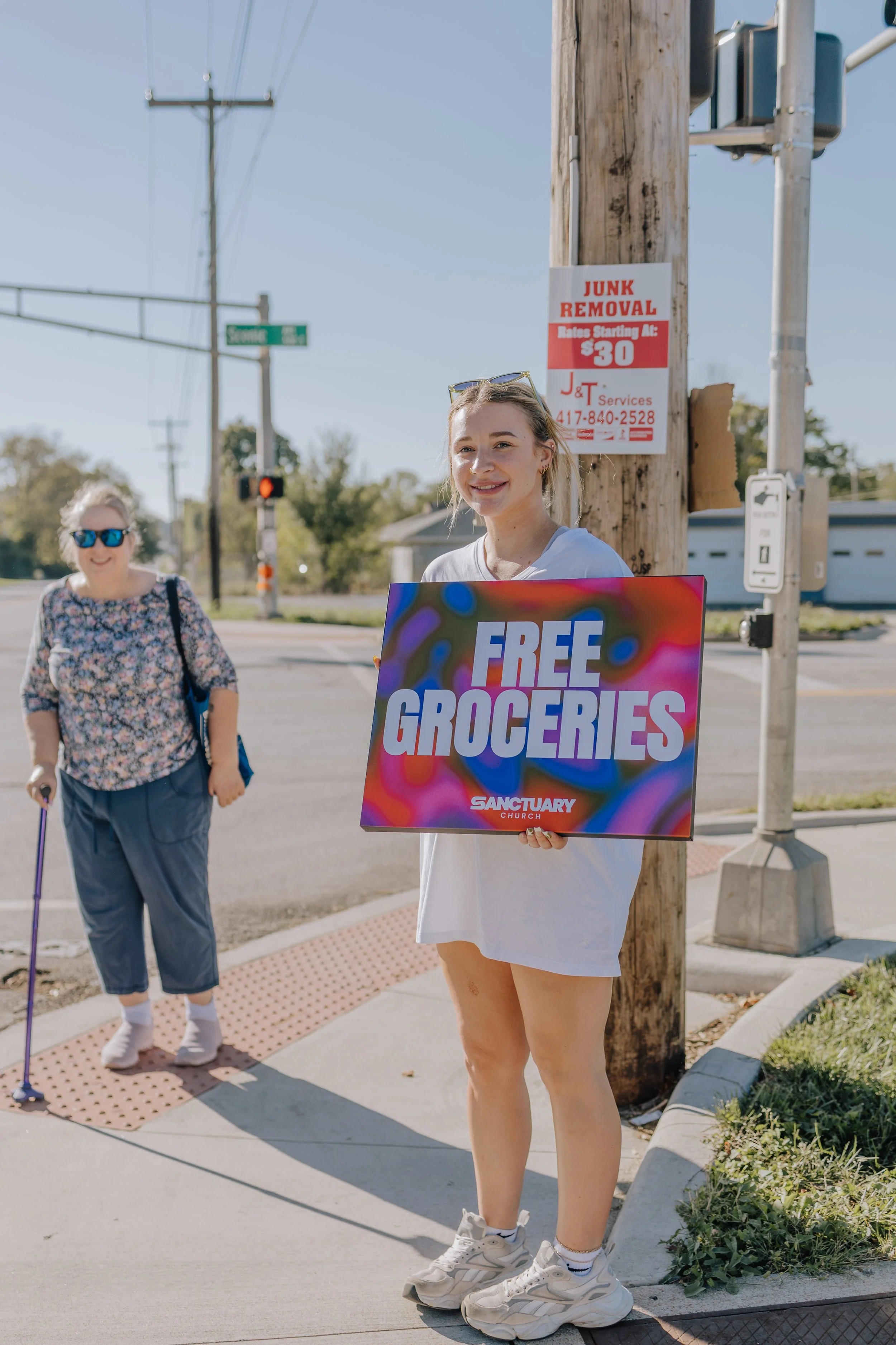 Young woman holding a colorful sign that says "FREE GROCERIES" on a sidewalk. Another person stands behind her, smiling, wearing sunglasses and a floral shirt.