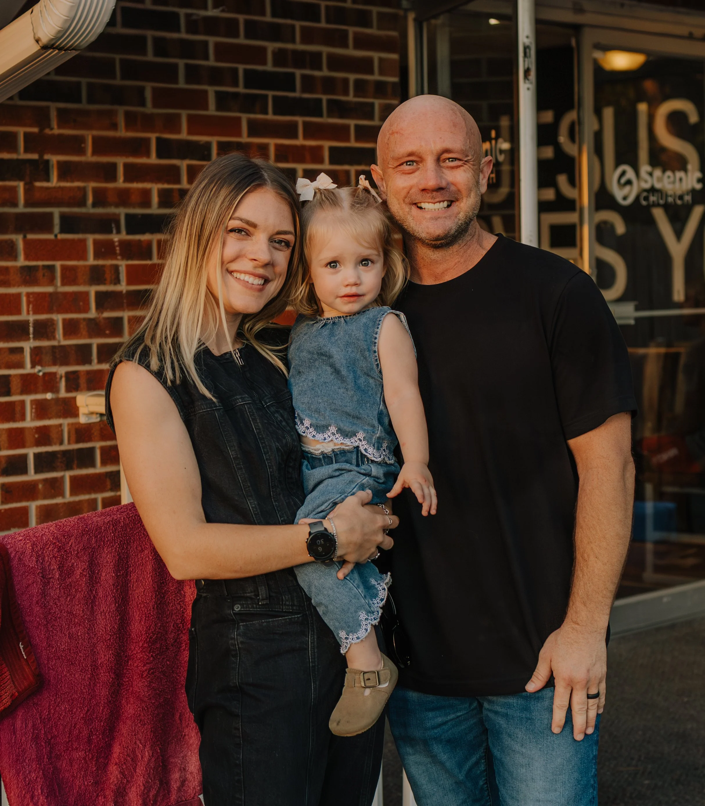 A smiling family of three standing in front of a brick wall, including a woman with blonde hair holding a young girl with blonde hair, and a man with a shaved head and beard. The young girl is looking at the camera while the woman and man smile.