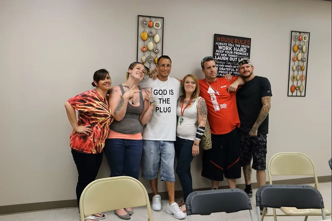 Six people standing in a room with plain walls, two decorative wall art pieces, and a sign with house rules. The group is smiling, with some posing and some leaning on each other.