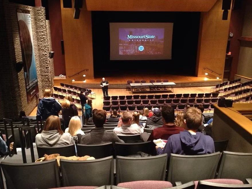 Audience seated in an auditorium with a speaker on stage, a large screen displaying "Missouri State University," and wooden theater walls.