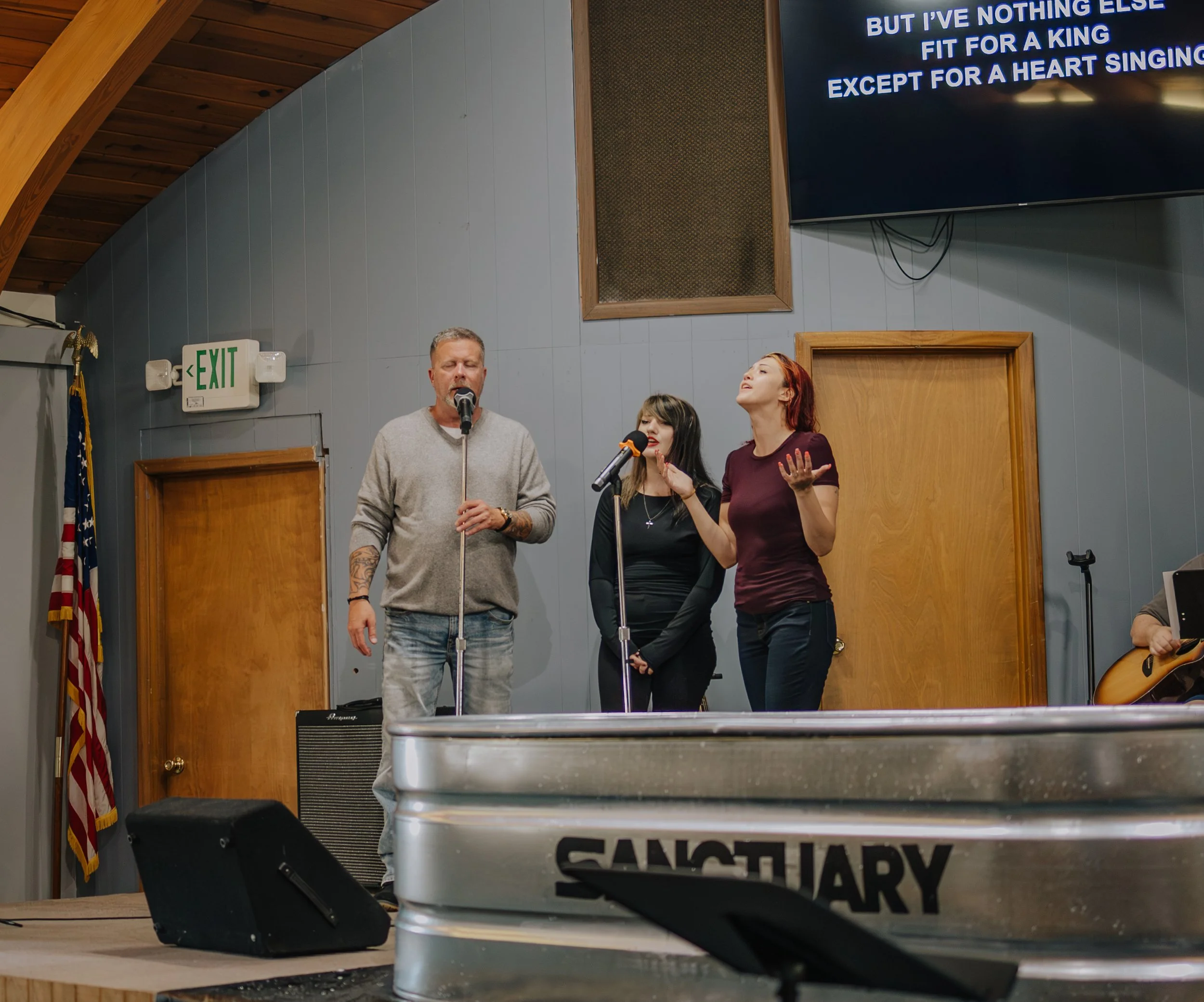 Three people singing on a church stage, with a man holding a microphone on the left and two women singing on the right, one with red hair and one with black hair. A guitar is partially visible on the far right. Behind them, a mounted screen displays song lyrics, an exit sign is on the wall, and an American flag is positioned next to a wooden door.