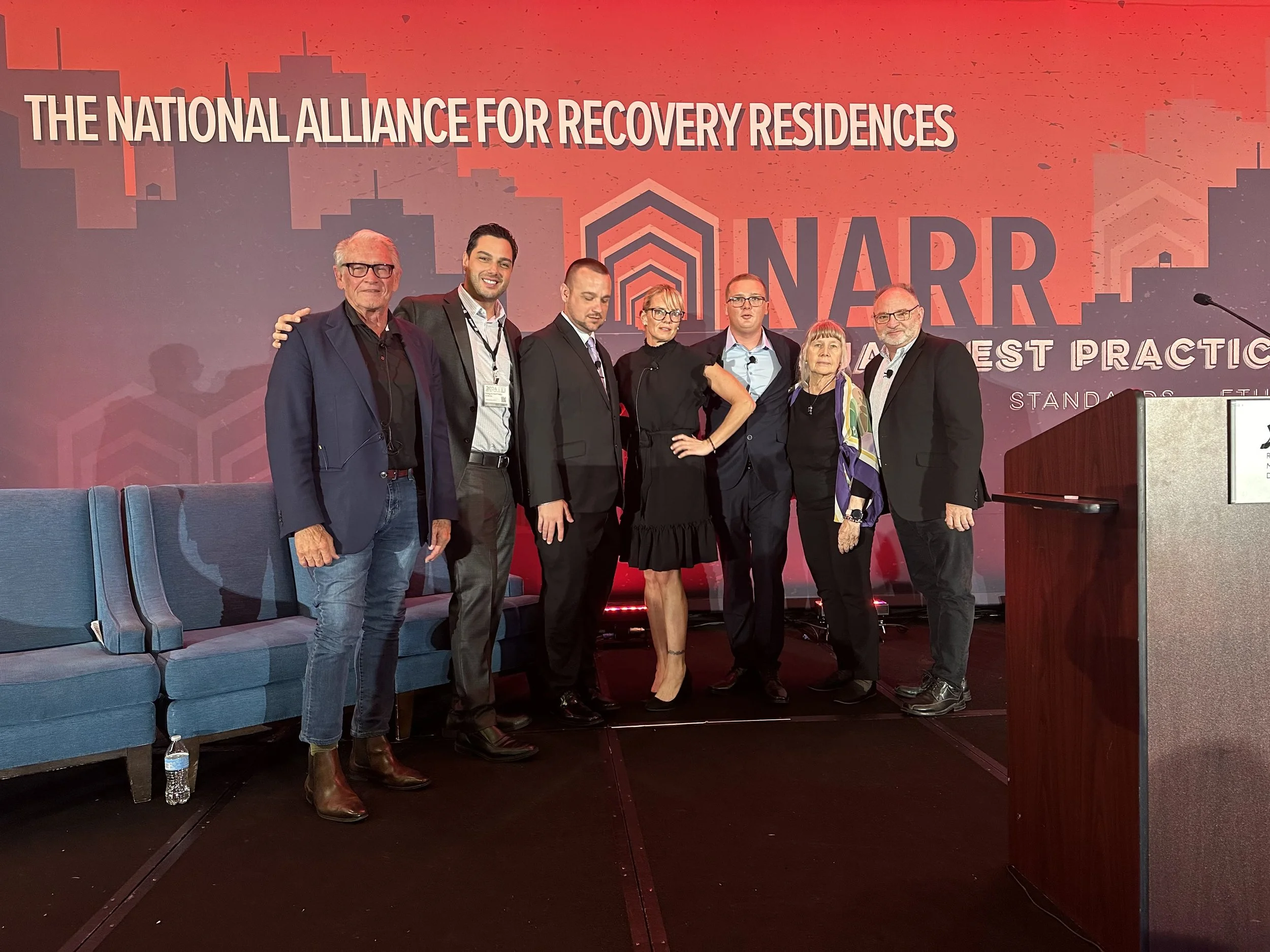 Group of seven people standing on stage at a conference with a background that reads 'The National Alliance for Recovery Residences' and 'NAARR'.