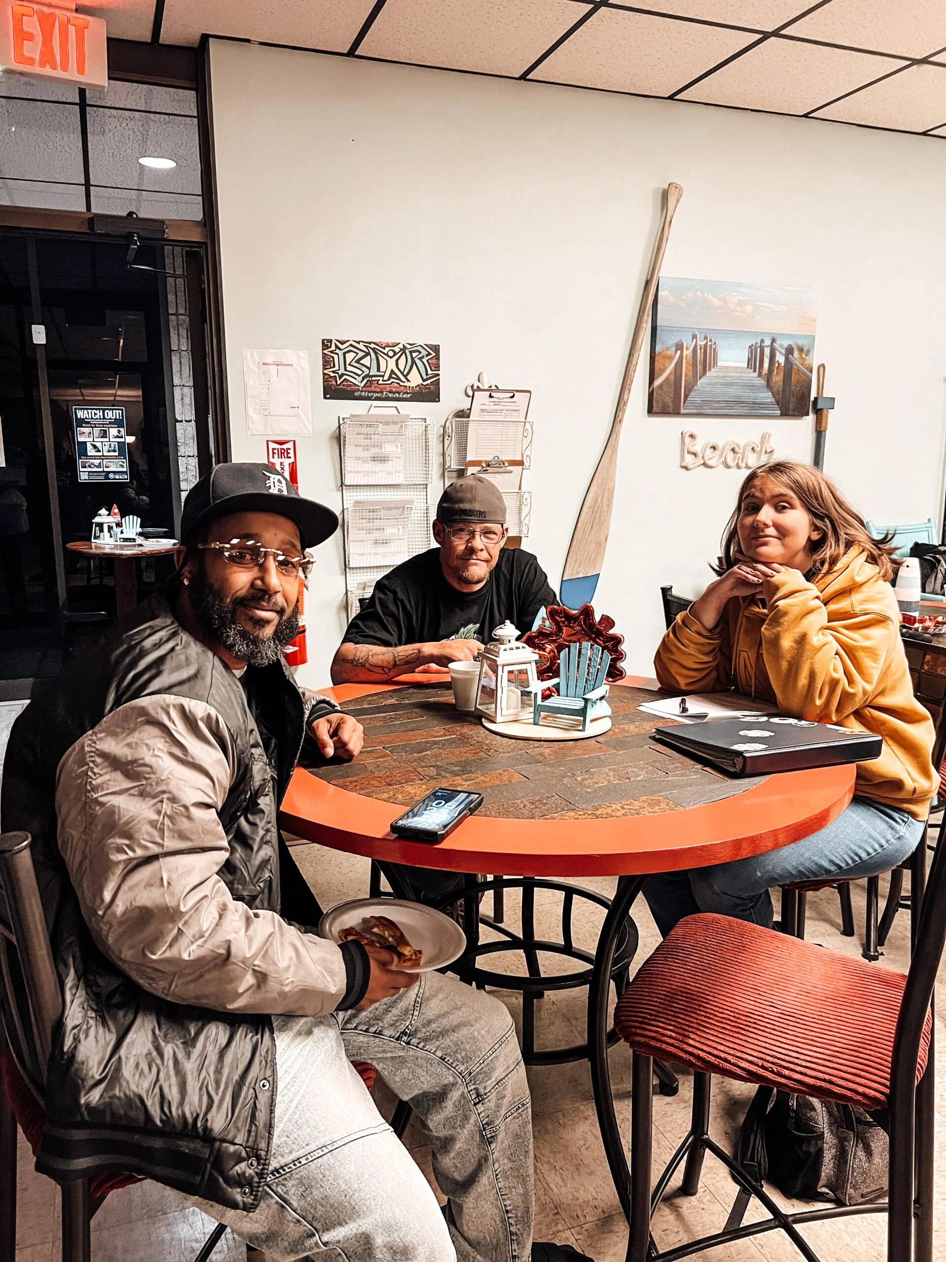 Three people sitting at a round table in a restaurant or café, with some decorations and a paddle in the background, and a menu trying to be seen. One man is holding a plate of food and has a phone on the table in front of him, a woman is sitting with her chin resting on her hands, and another man is sitting behind the table with a coffee cup in front of him.