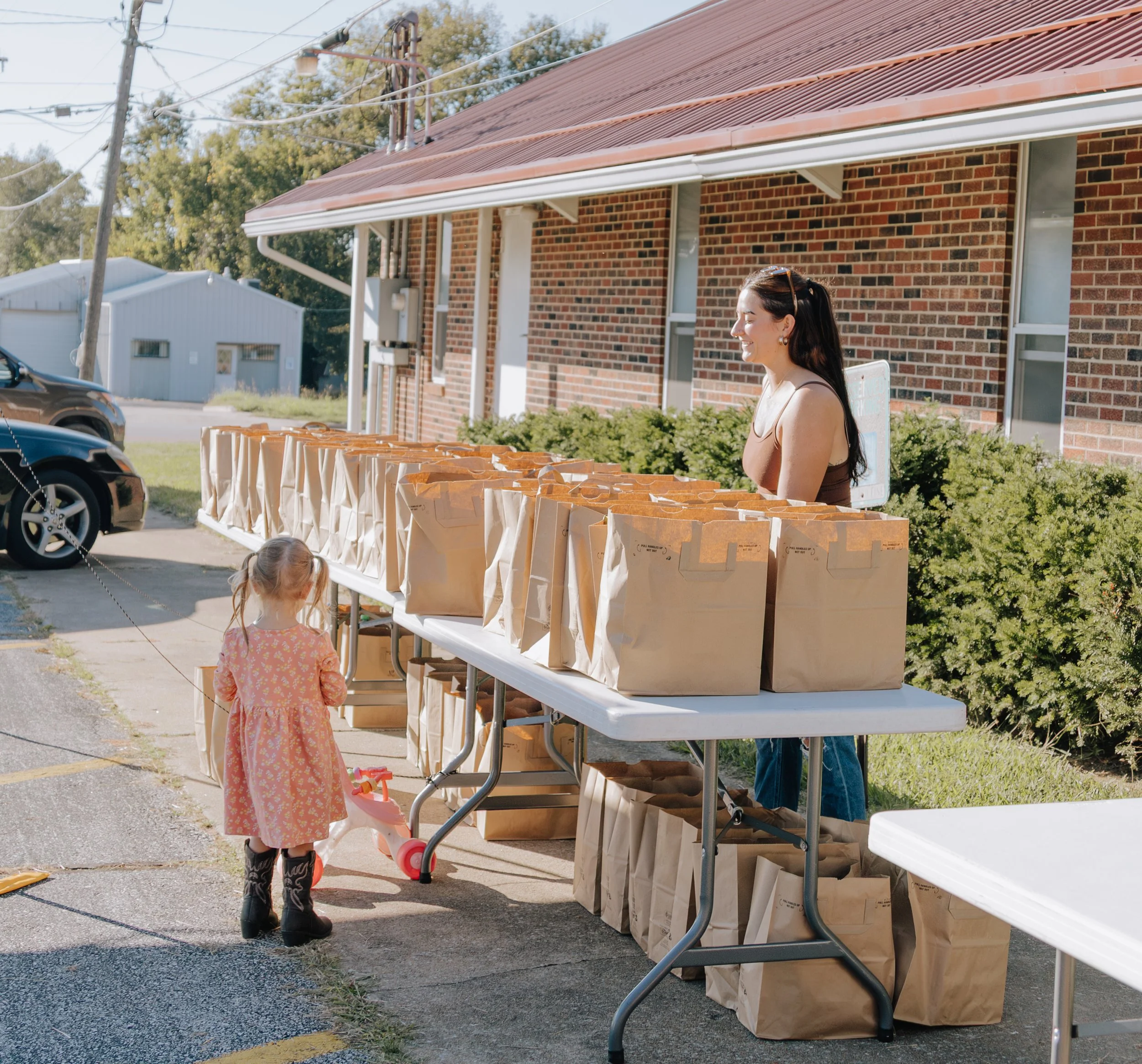 A woman and a young girl at an outdoor table with paper bags, possibly at a community event or charity distribution.