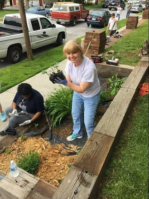 A woman smiling and holding a potted plant in a garden bed, with another person working nearby, during a gardening activity on a sidewalk near parked cars and a brick wall.