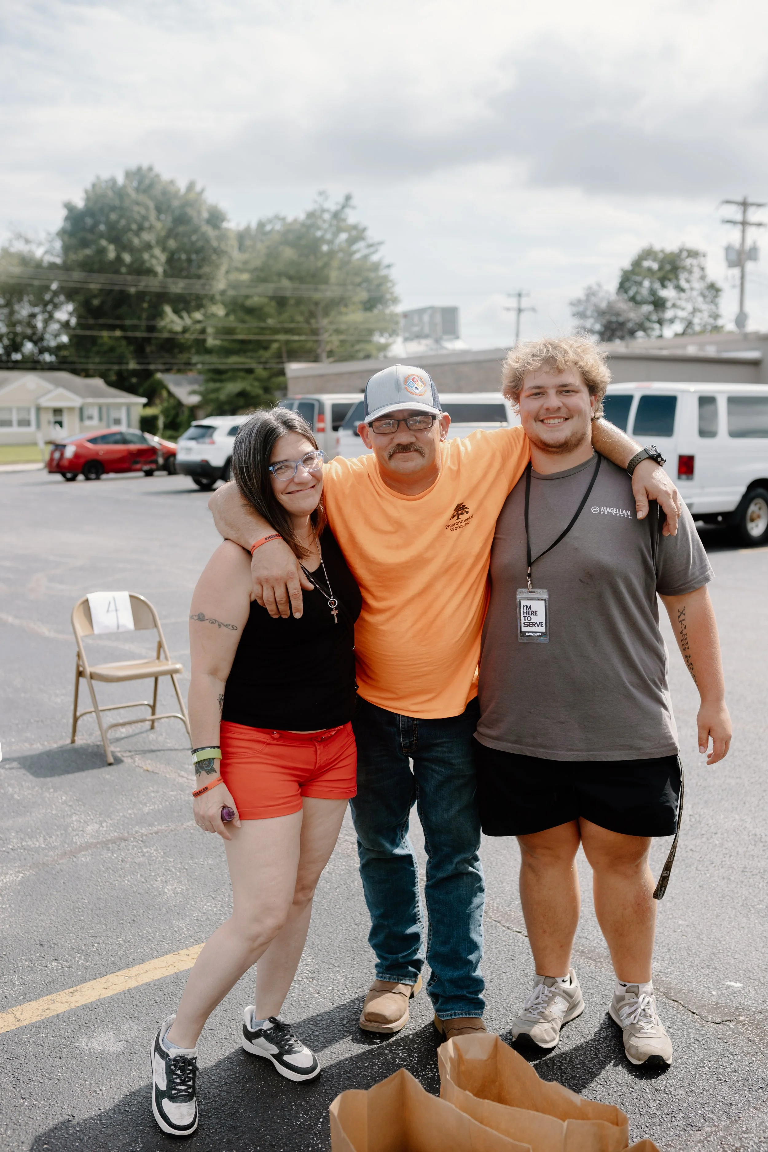 Three people standing together in a parking lot, smiling, with cars and trees in the background. They have their arms around each other.