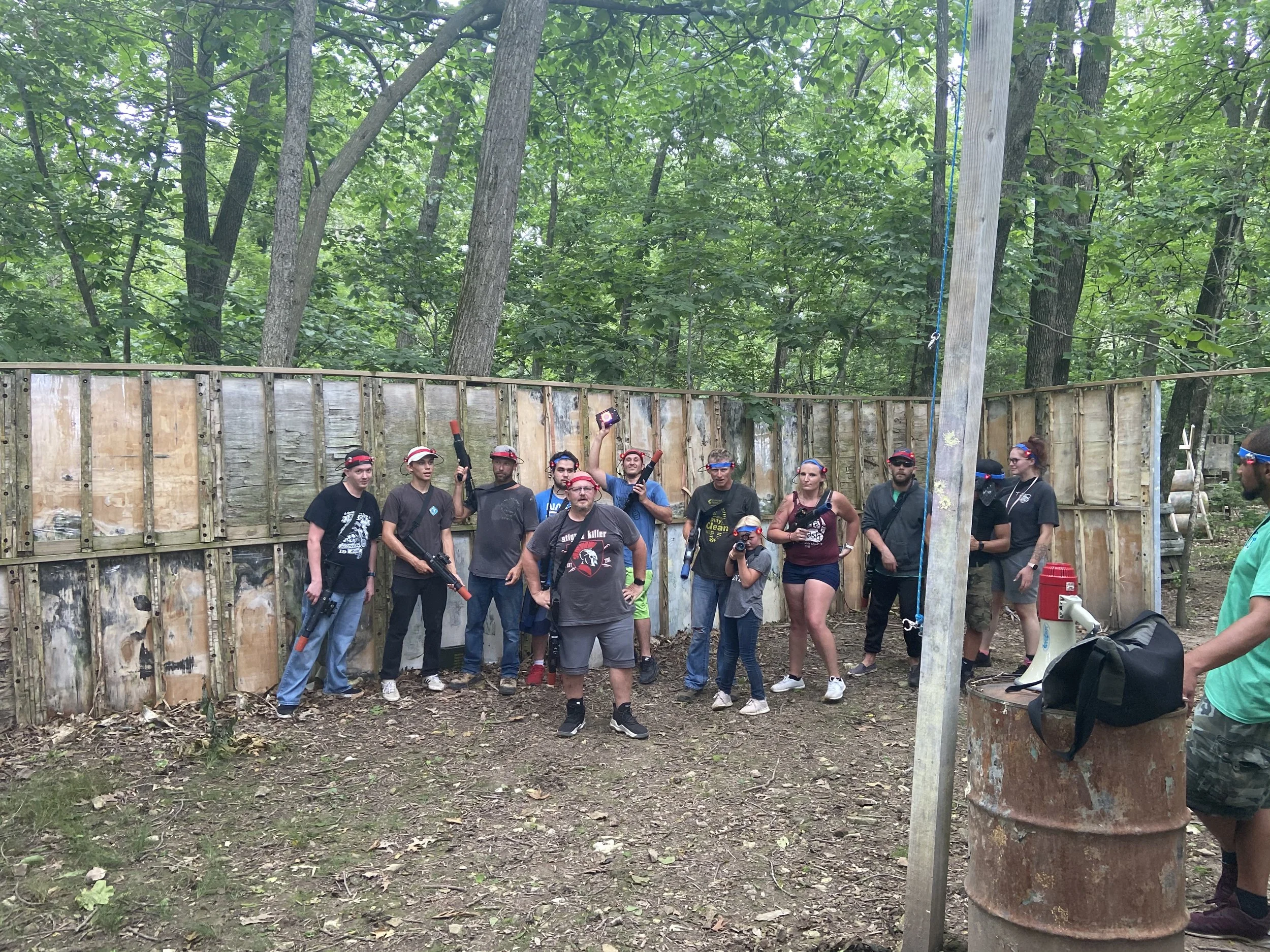 Group of people at outdoor laser tag arena standing in front of wooden fence with trees in the background. Some are holding laser guns, wearing goggles, and a person is taking a photo with a phone. There is a rusty barrel with a megaphone and a bag on top, and a person on the right side wearing a green shirt.