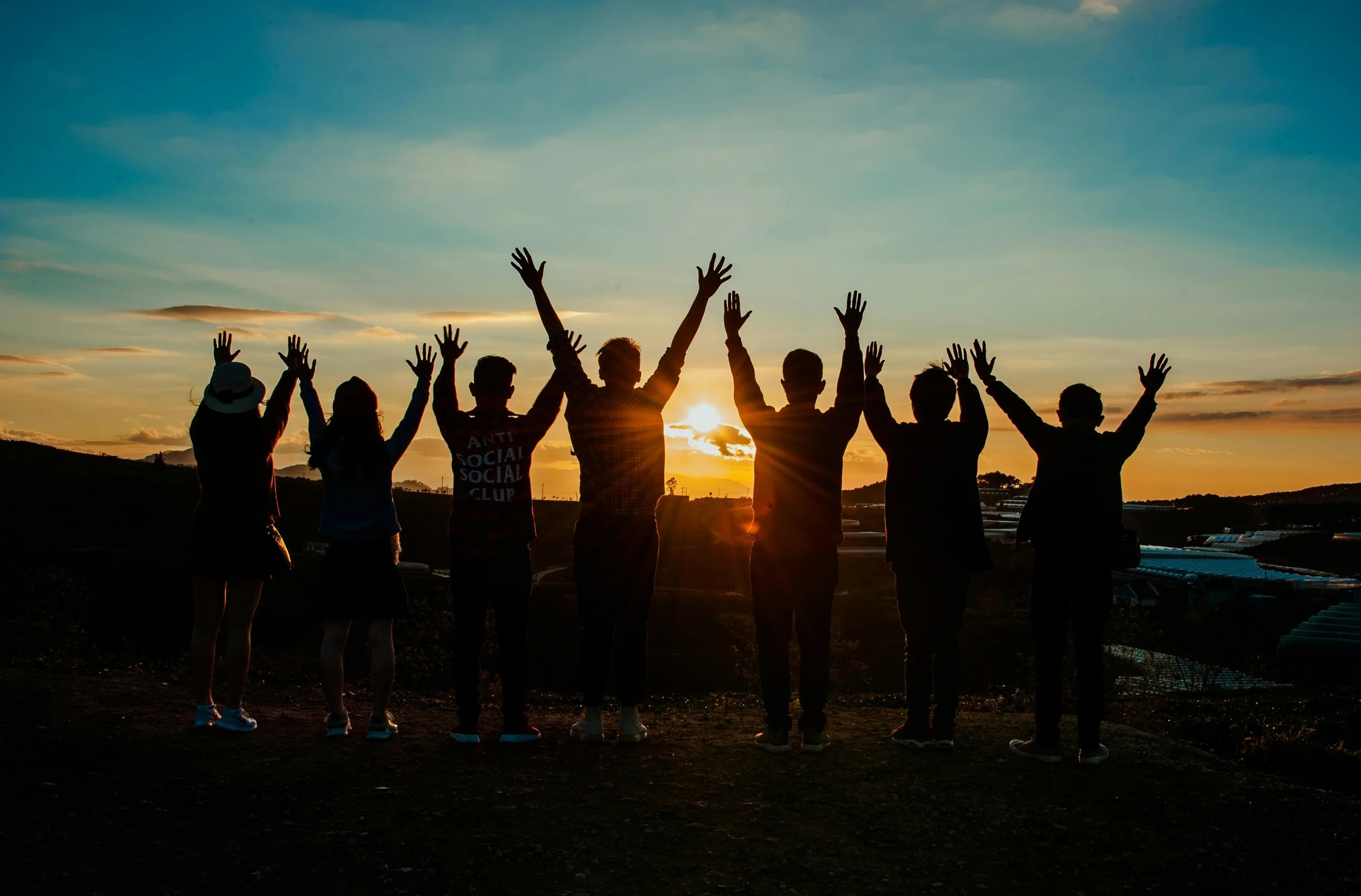 Group of seven people standing outdoors at sunset with their arms raised in the air, silhouetted against the sky and setting sun.