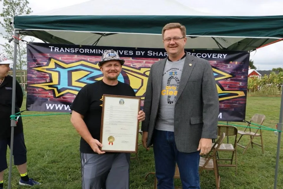 Two men are standing outdoors in front of a tent with a banner. One man is holding a framed certificate, smiling, and wearing a hat and t-shirt. The other man is taller, wearing glasses, a gray blazer, and a t-shirt. There are chairs behind them on the grass, and a person partially visible on the left.