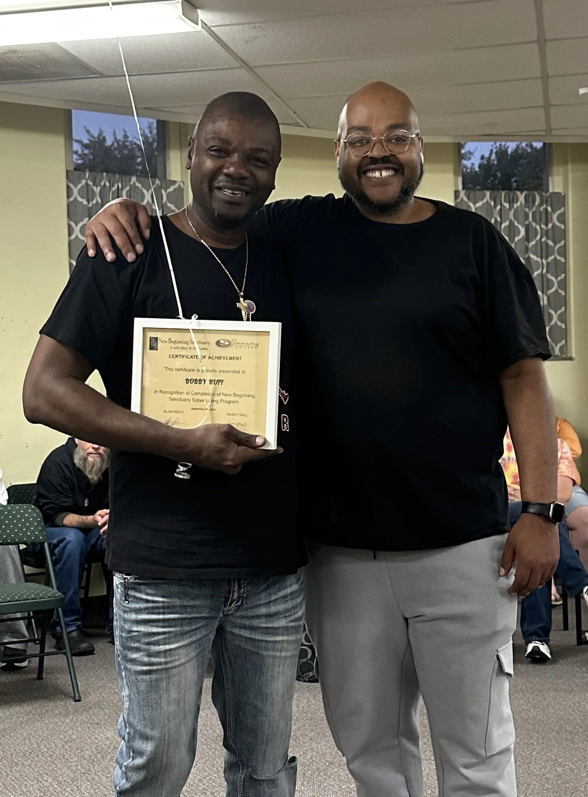 Two men standing side by side in a room. The man on the left is holding a framed certificate of achievement and has his arm around the shoulder of the man on the right. Both are smiling. Several other people are seated in the background.